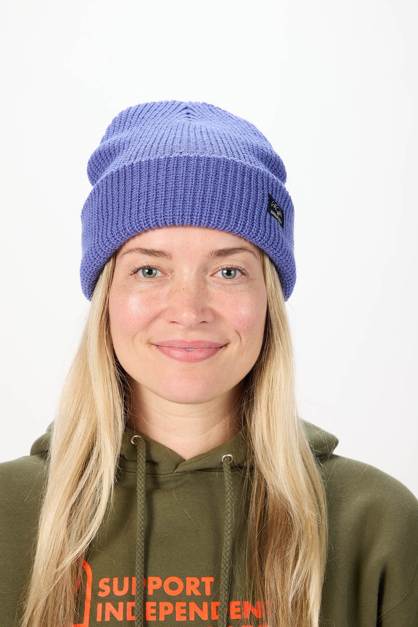A woman with long blonde hair wearing a purple ribbed knit beanie and an olive green hoodie with orange text, photographed against a plain white background.