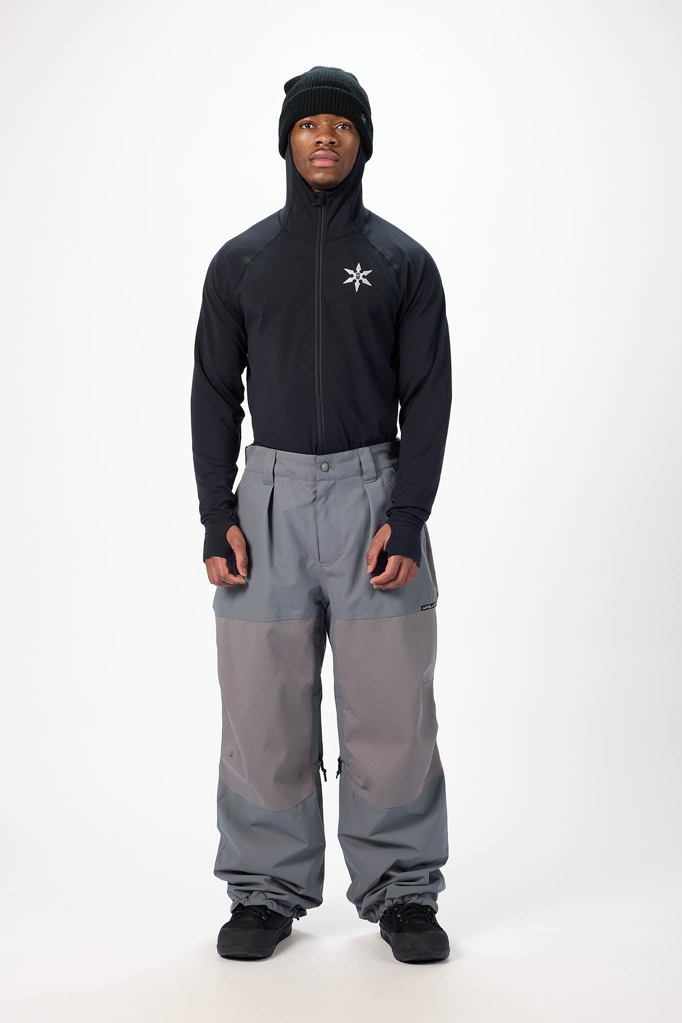 Person wearing a black jacket with a logo and gray pants on a white background
