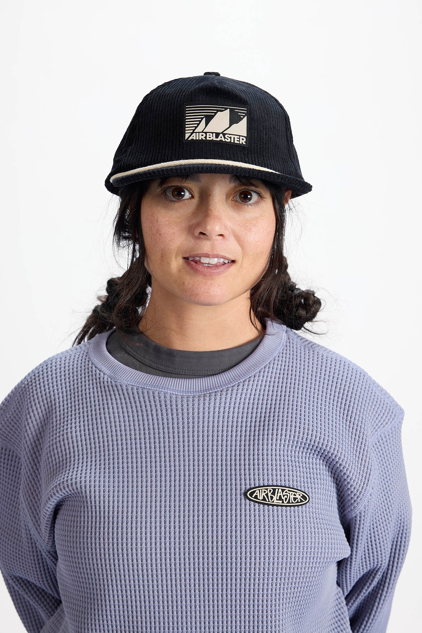 A woman wearing a black Airblaster baseball cap and a lavender waffle-knit sweatshirt with a small Airblaster logo on the chest, photographed against a plain white background.