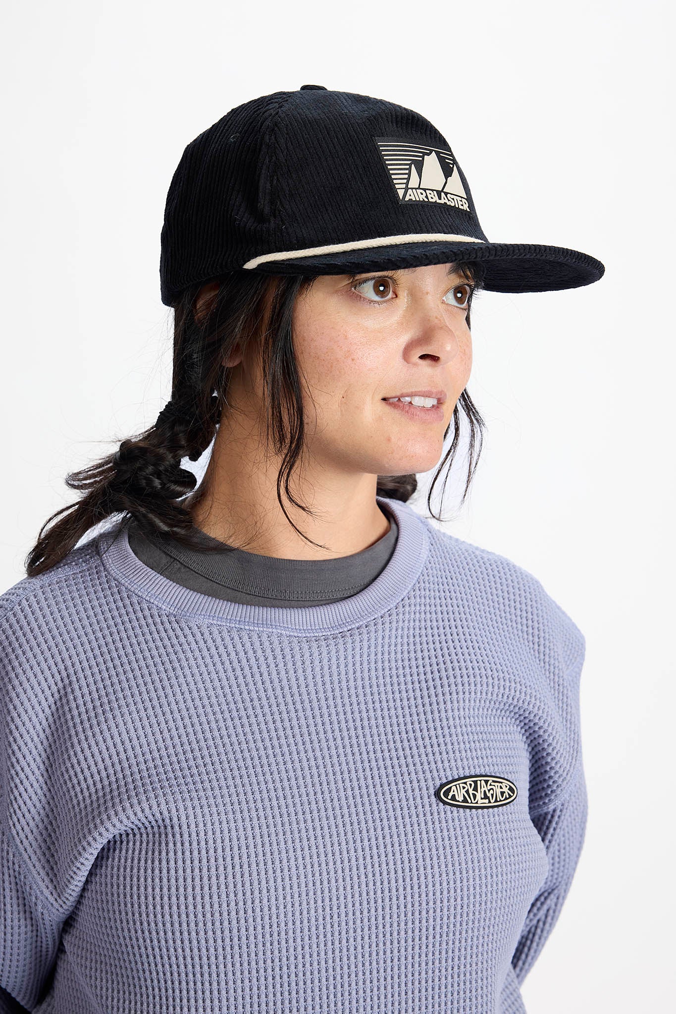 A woman wearing a black Airblaster baseball cap and a lavender waffle-knit sweatshirt with a small Airblaster logo on the chest, photographed against a plain white background.