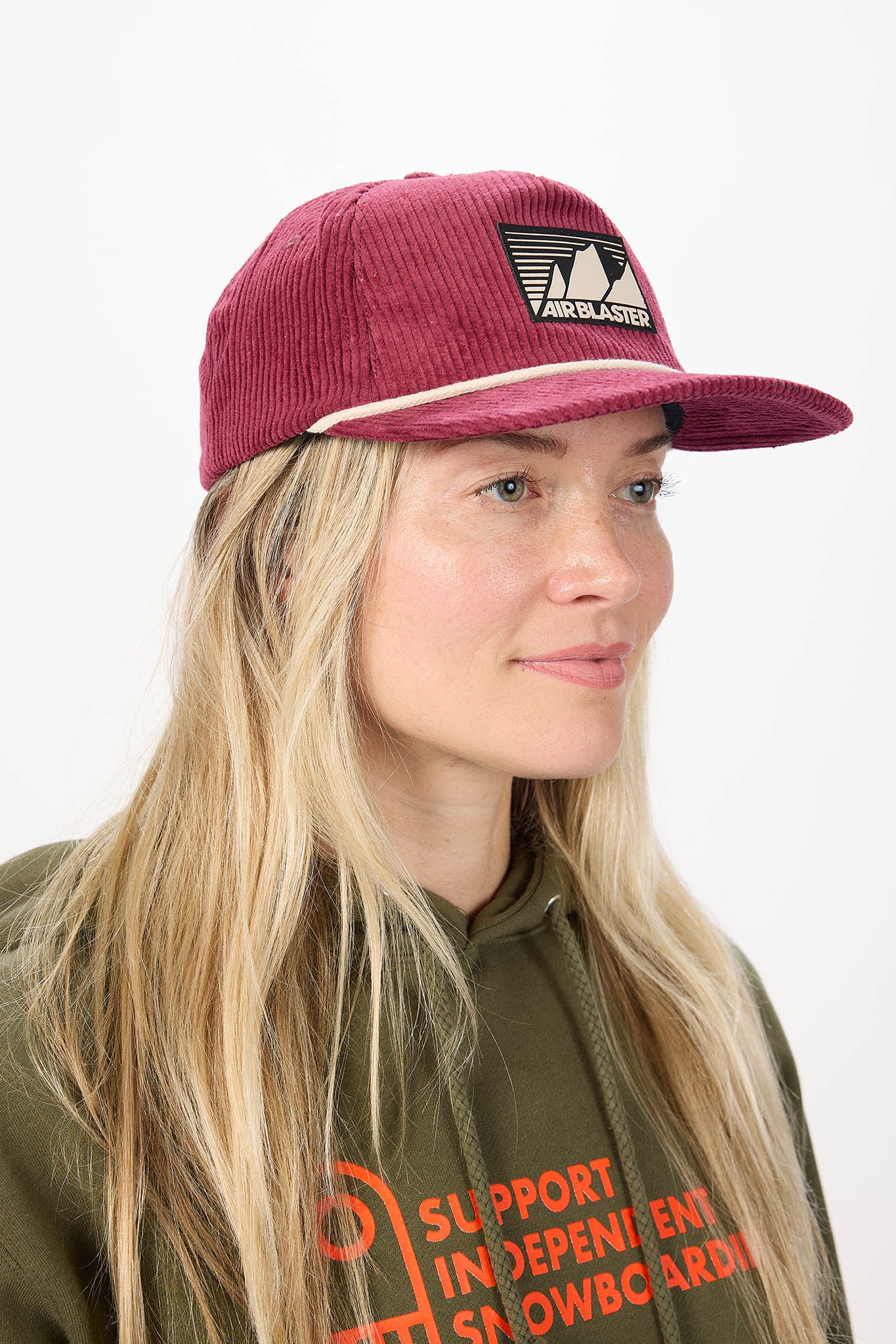A woman with long blonde hair wearing a burgundy corduroy cap with a black and white logo patch