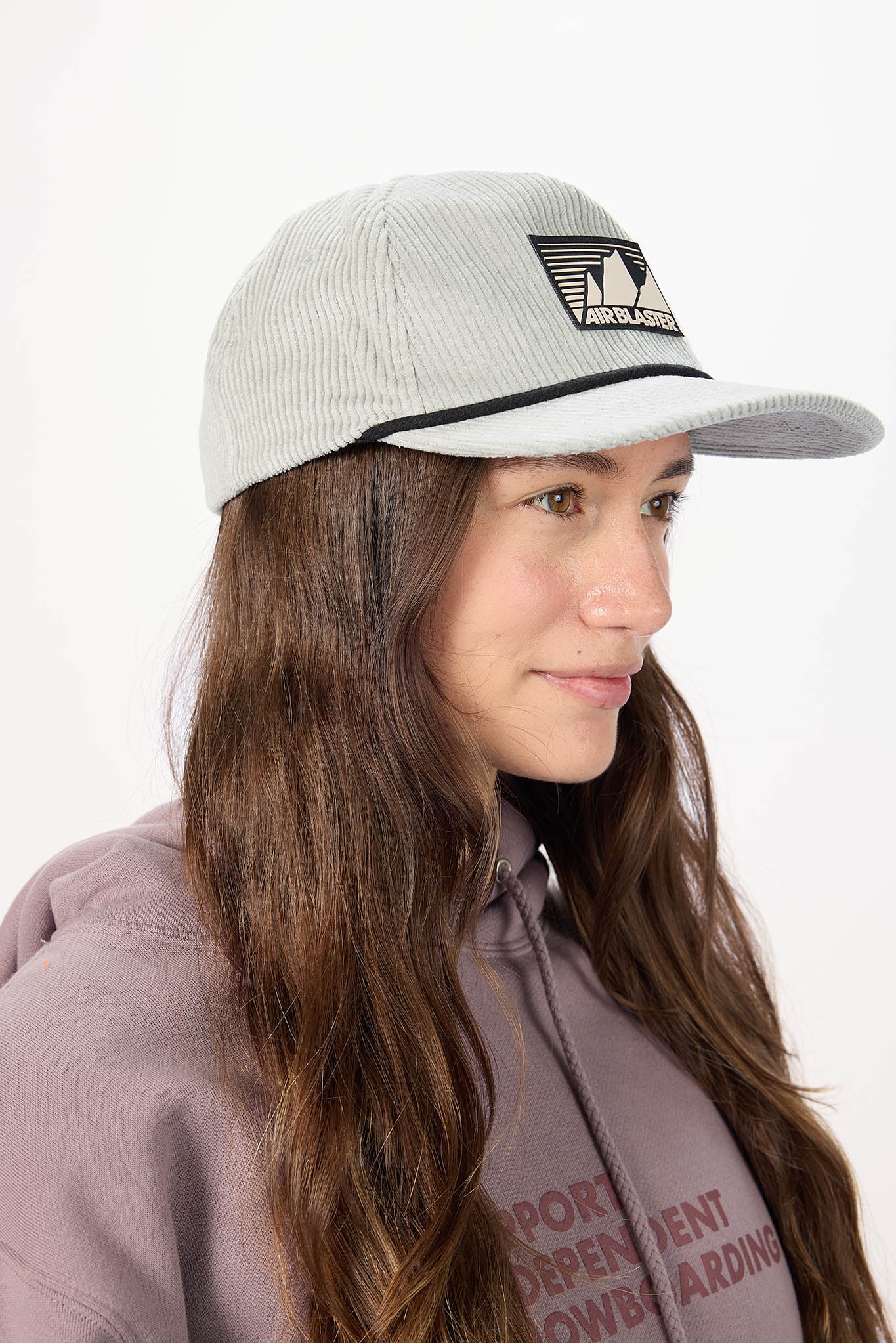 A woman wearing a light gray corduroy baseball cap with a black logo patch on the front, against a plain white background.