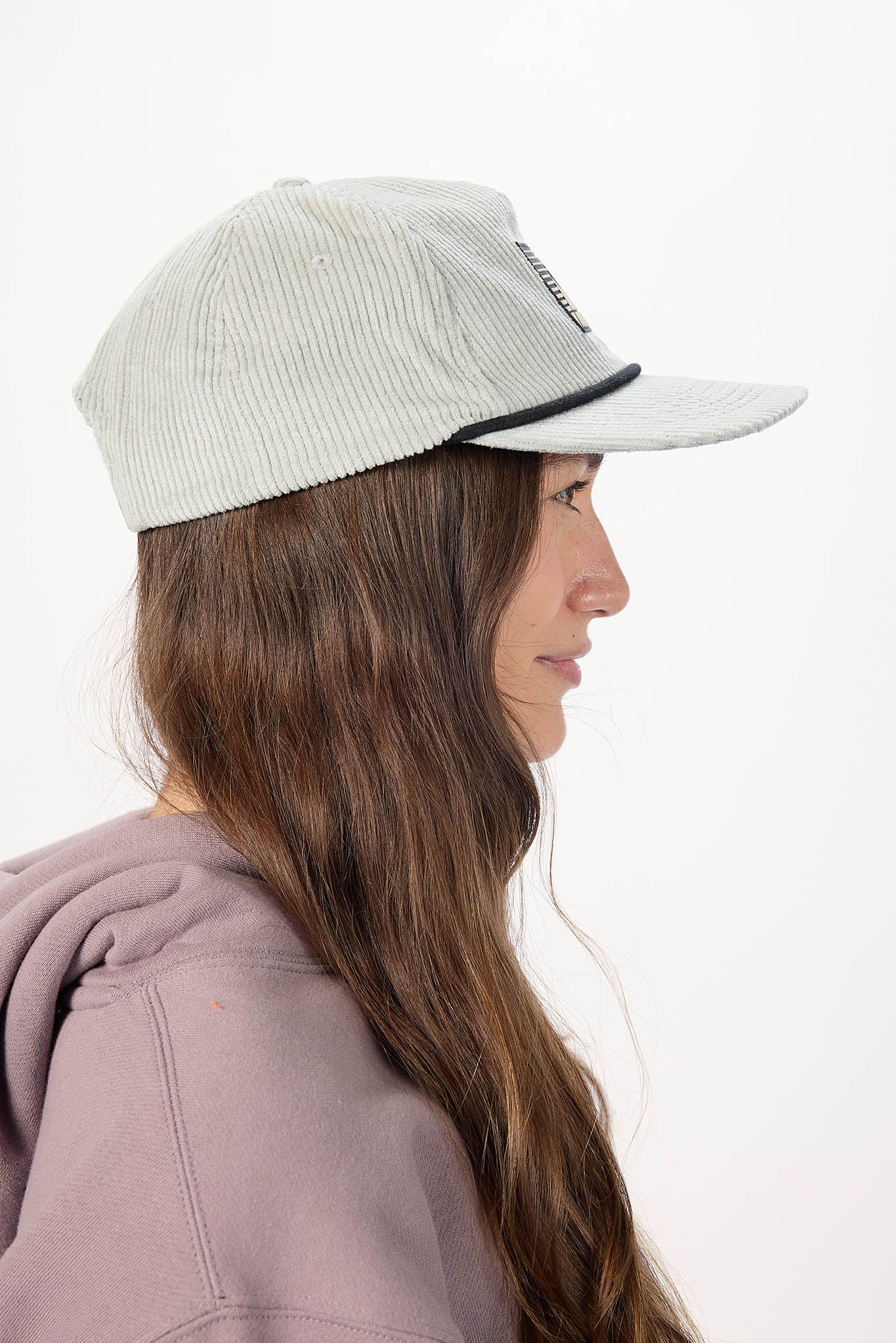A woman in profile wearing a light gray ribbed corduroy cap with a black trim detail on the brim, paired with a dusty purple hoodie, against a plain white background.