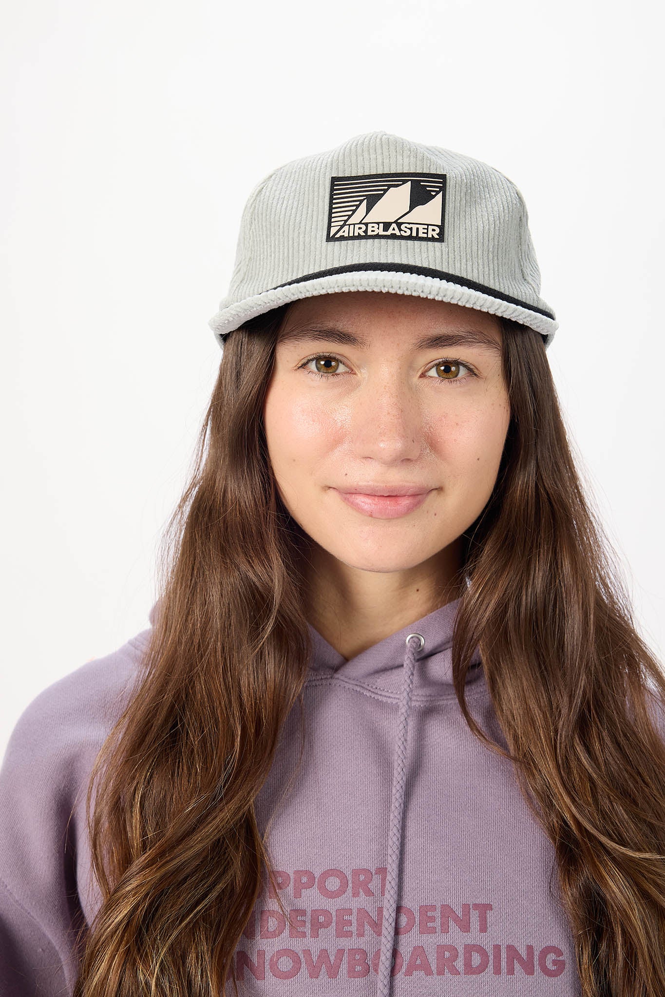 A woman with long brown hair wearing a light gray ribbed knit cap with a black Airblaster logo patch, paired with a lavender hoodie against a plain white background.