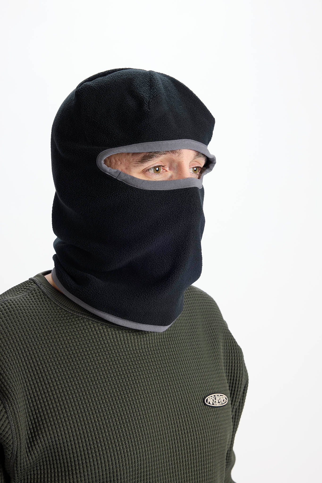 A person wearing a black balaclava face covering with gray trim, paired with an olive green waffle-knit shirt against a plain white background.