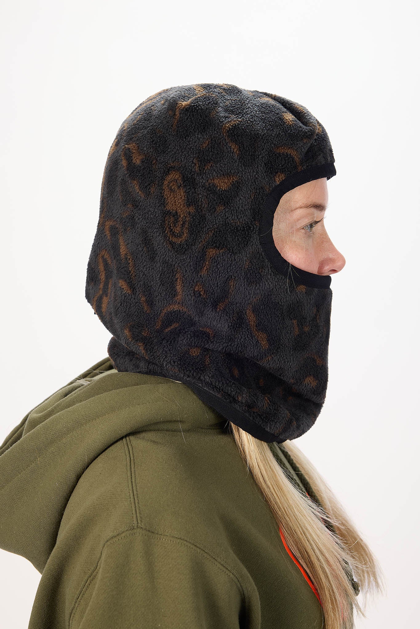 A woman wearing a black fleece balaclava mask with a brown leopard print pattern, paired with an olive green hoodie against a plain white background.