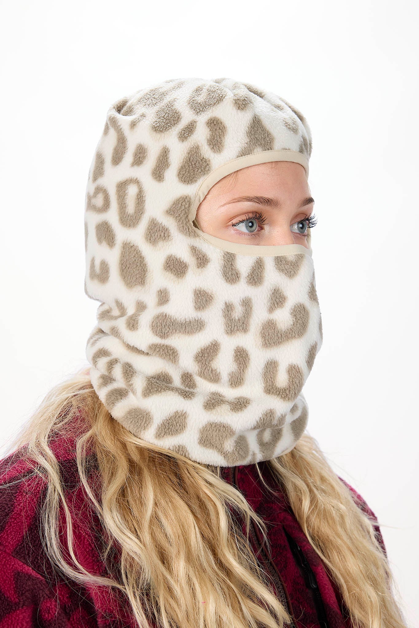 A woman wearing a white leopard patterned balaclava mask with a brown leopard print pattern, paired with an olive green hoodie against a plain white background.