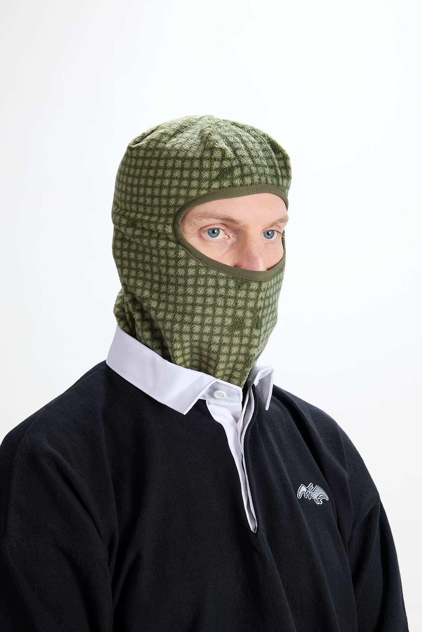 A man wearing an olive green patterned balaclava face covering over a black long-sleeve shirt with a white collar, photographed against a plain white background.