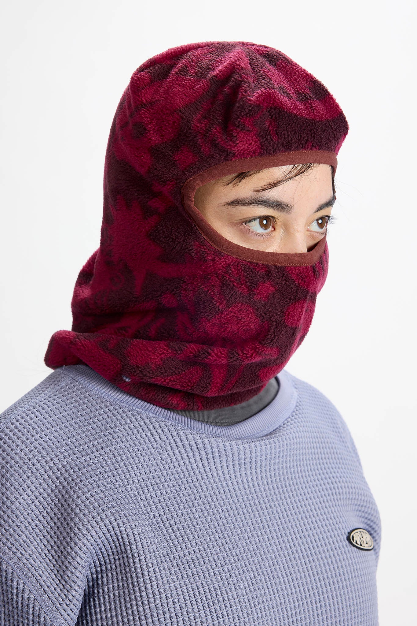 A person wearing a burgundy and black patterned fleece balaclava hood over a light purple waffle-knit sweater, shown against a plain white background.