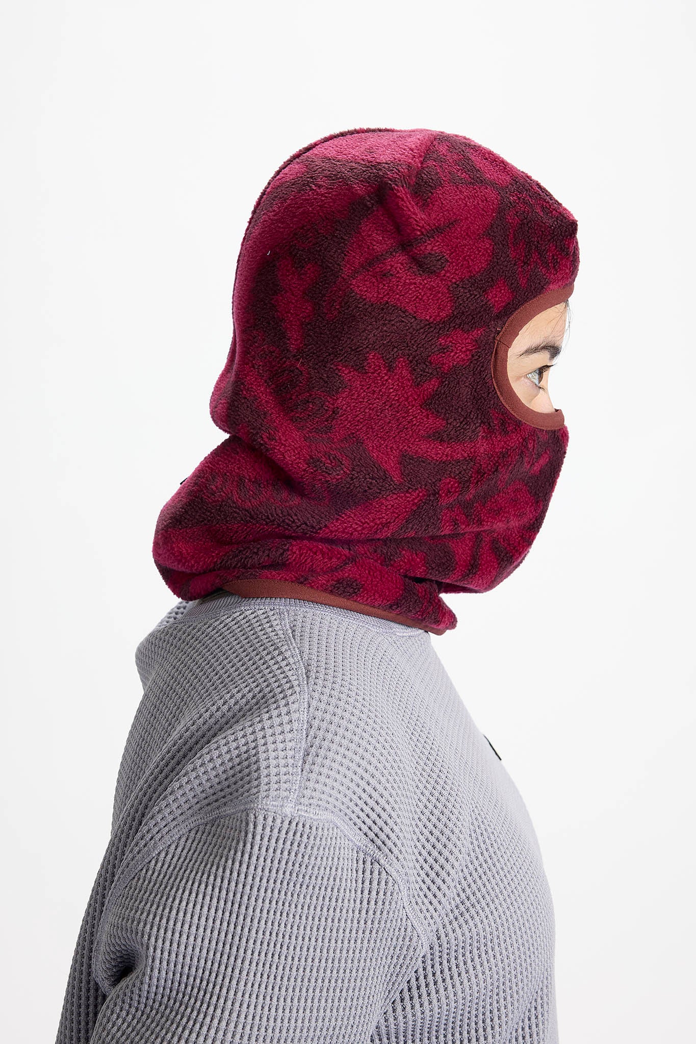 A side profile of a person wearing a burgundy and dark red patterned fleece balaclava mask over a gray waffle-knit sweater, set against a plain white background.