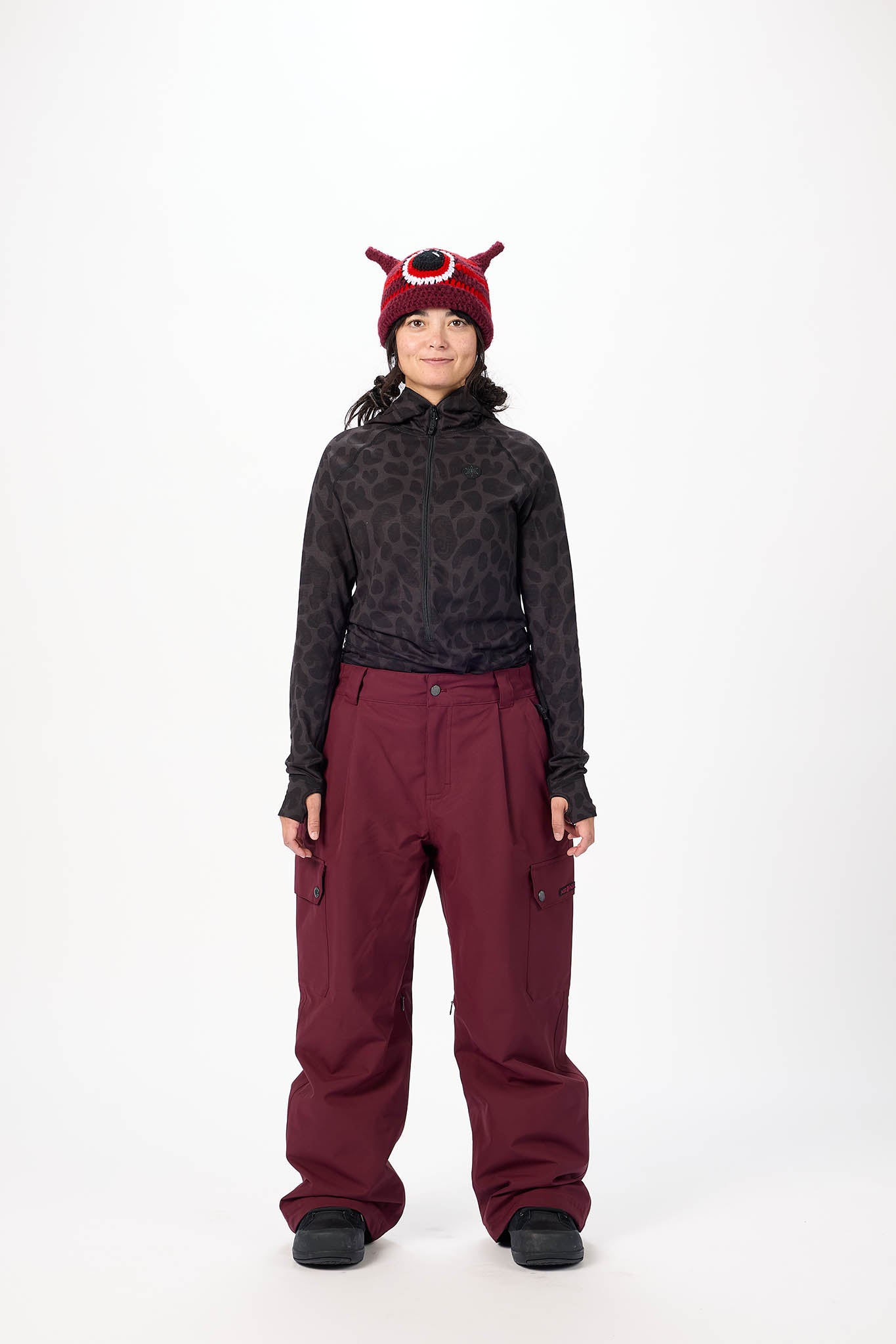 Person wearing a dark patterned long-sleeve top and maroon pants with a red hat on a white background