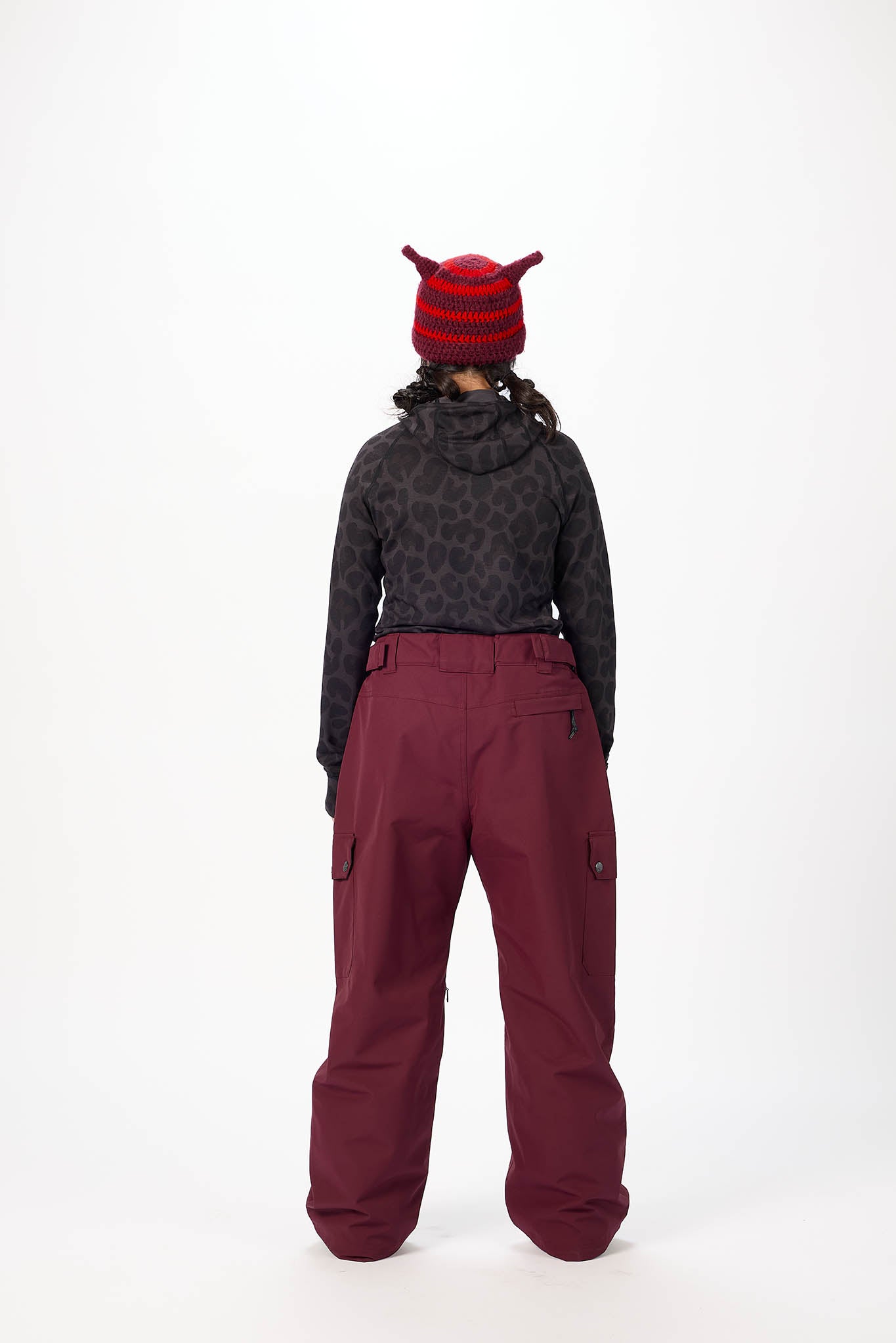 Person wearing a dark patterned jacket and maroon pants with a red and black beanie on a white background.
