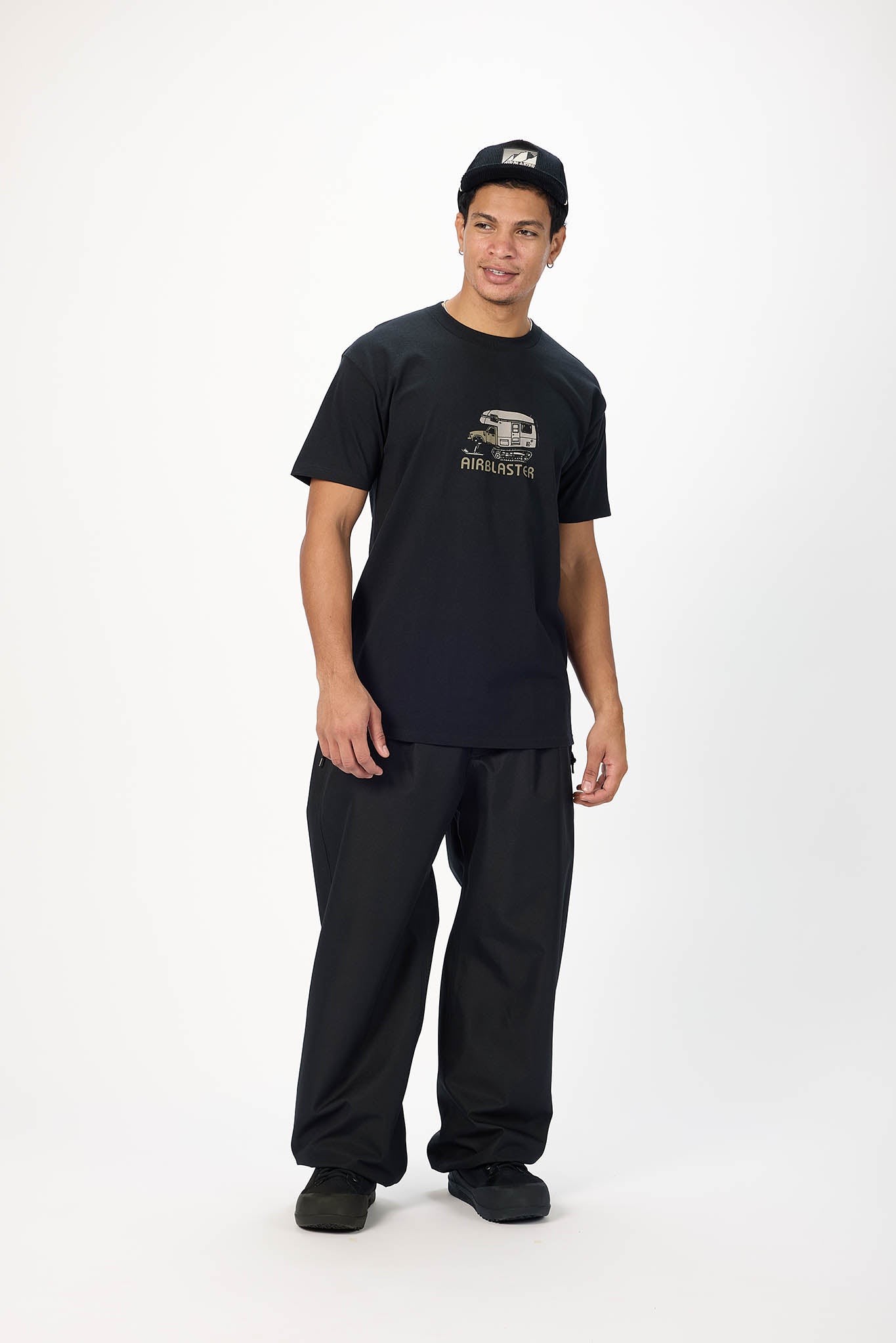 A man modeling a black crewneck t-shirt with a gold graphic on the chest, paired with black sweatpants and a black cap, standing against a plain white studio background.