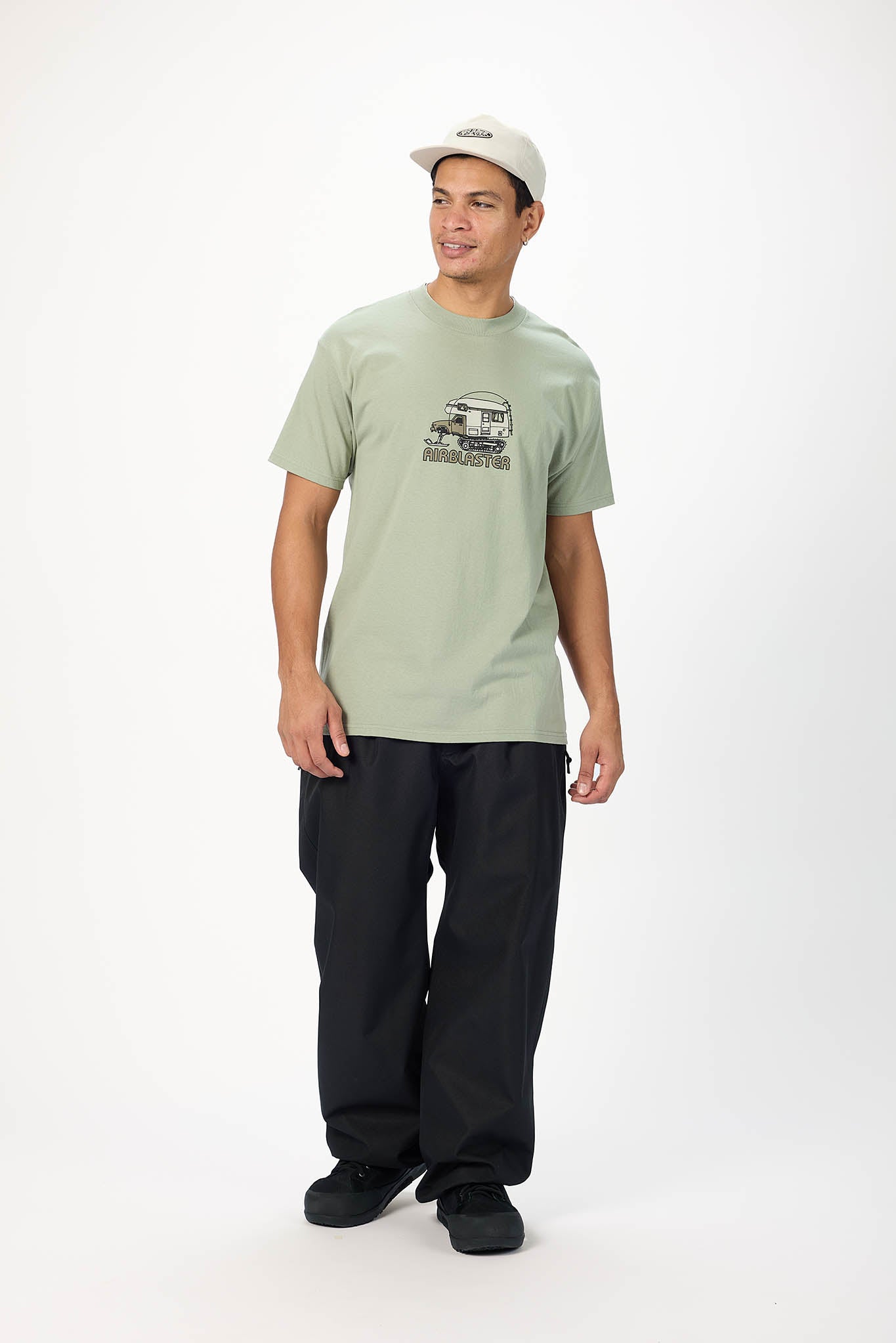 A man modeling a light sage green t-shirt with a black graphic print on the chest, paired with black pants and a white baseball cap, against a plain white studio background.