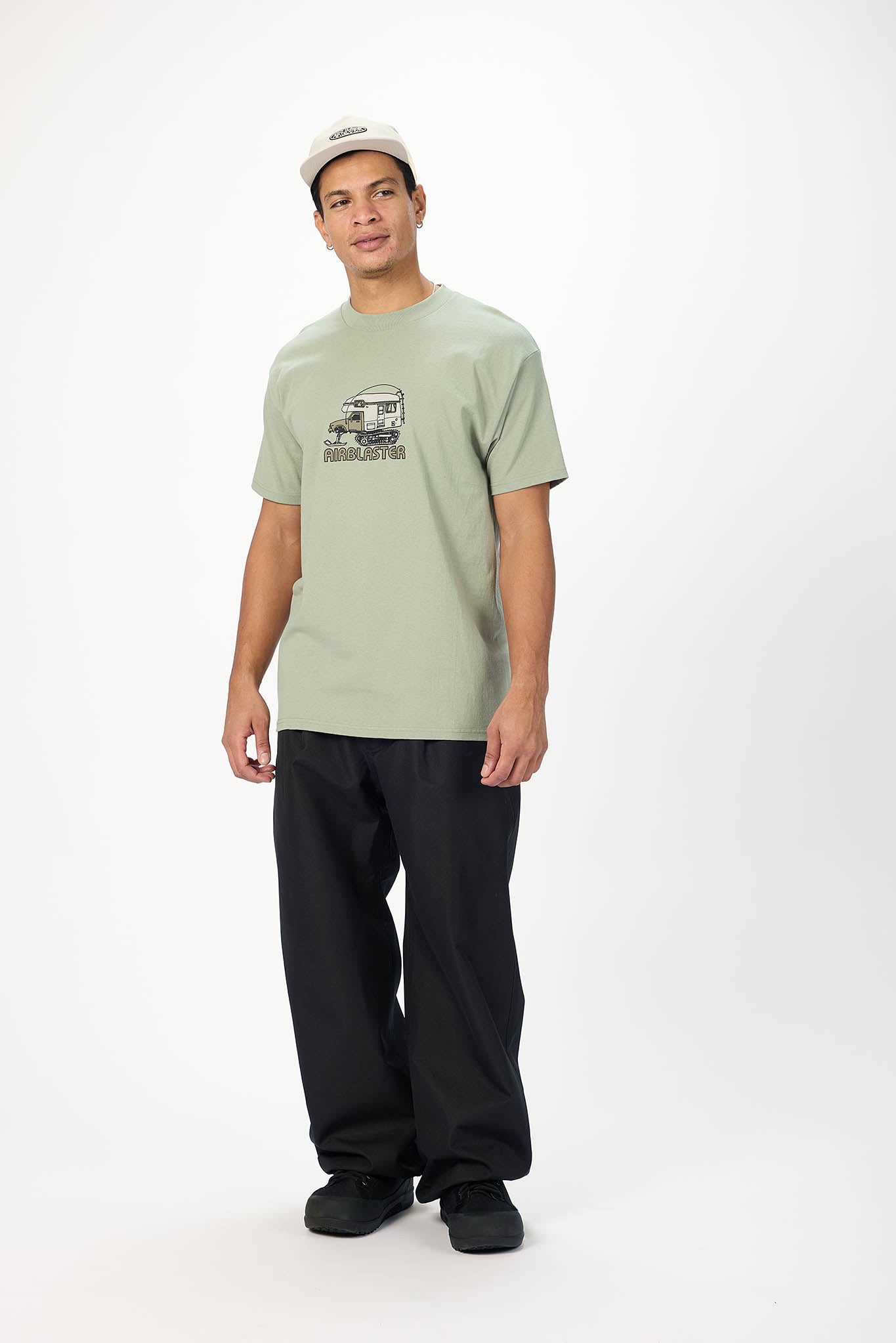 A man modeling a light sage green t-shirt with a black graphic of a vintage camper and the text 'CAMPERS' on the chest, paired with black pants and a beige baseball cap, against a plain white studio background.