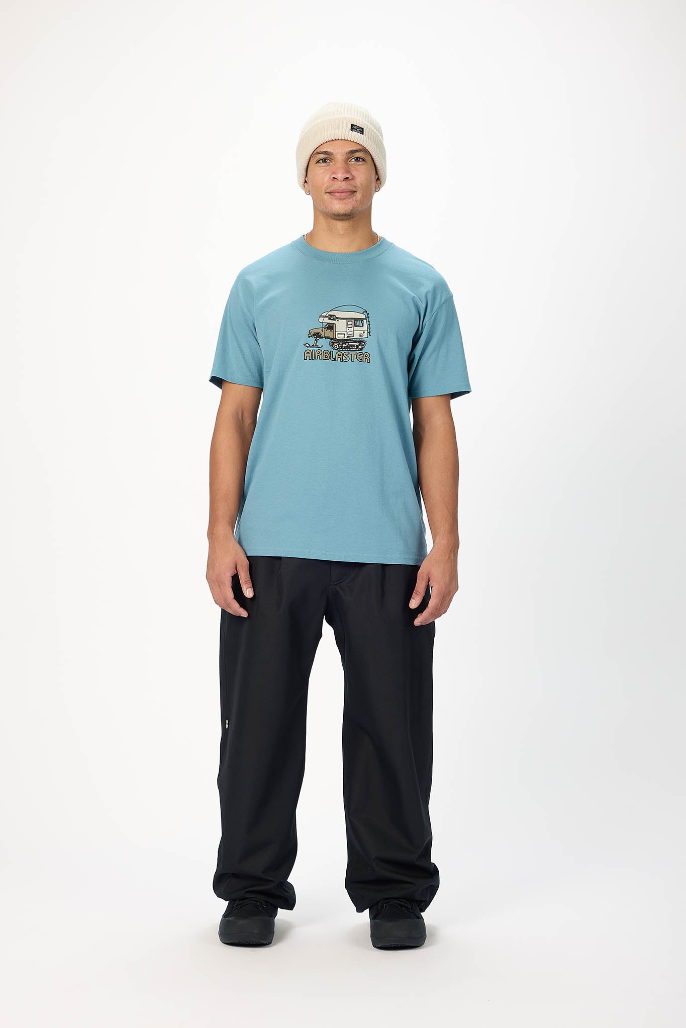 A man modeling a light blue crewneck t-shirt with a vintage camper graphic on the chest, paired with black sweatpants and a beige beanie, standing against a plain white studio background.