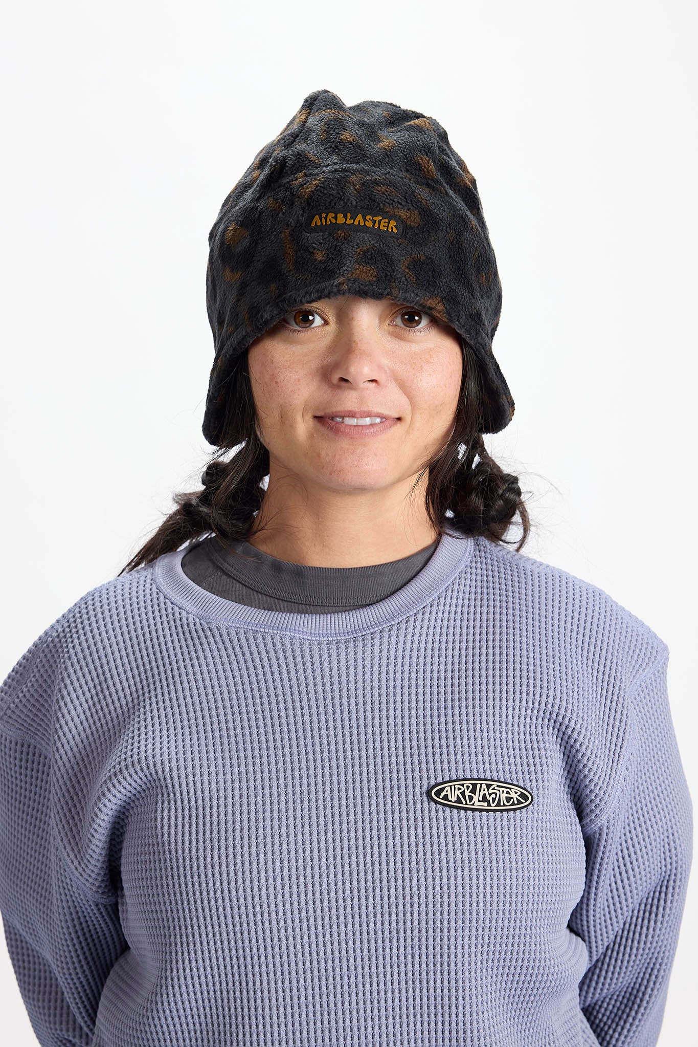 A woman wearing a black leopard print Airblaster beanie and a lavender waffle-knit crewneck sweatshirt with an AirBlaster logo on the chest, photographed against a plain white background.