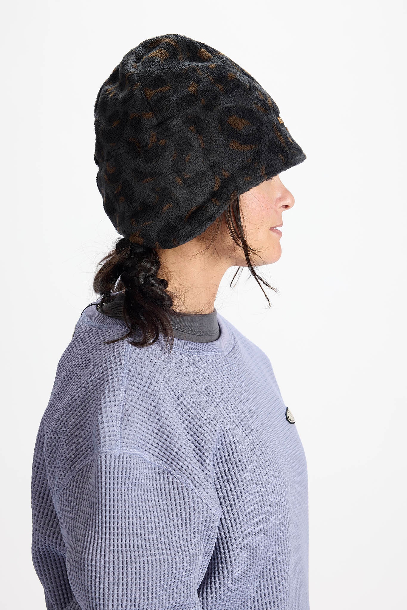 A side-profile view of a person wearing a dark brown and black leopard-print fuzzy beanie and a textured lavender-gray waffle-knit sweatshirt against a plain white background.