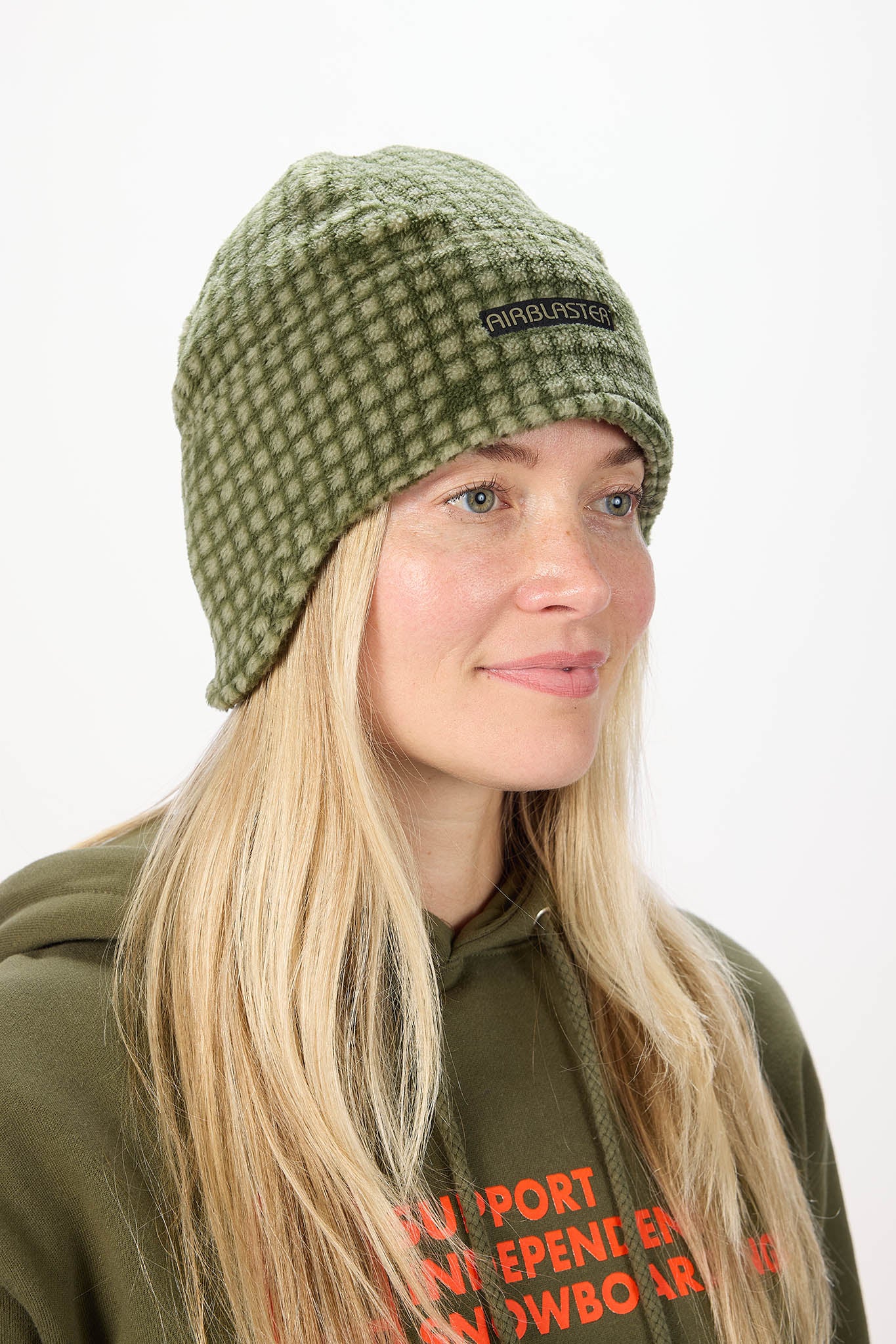 A woman wearing an olive green and white checkered knit beanie with a black brand patch that reads 'AirBlaster'. She is also wearing an olive green hoodie with orange text.