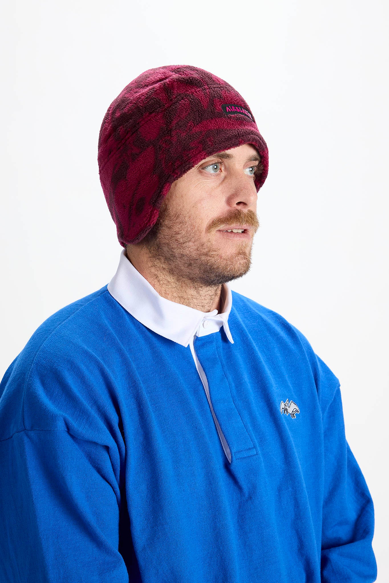 A man wearing a burgundy and black patterned knit beanie and a blue rugby-style pullover sweater over a white collared shirt, photographed against a plain light gray studio background.