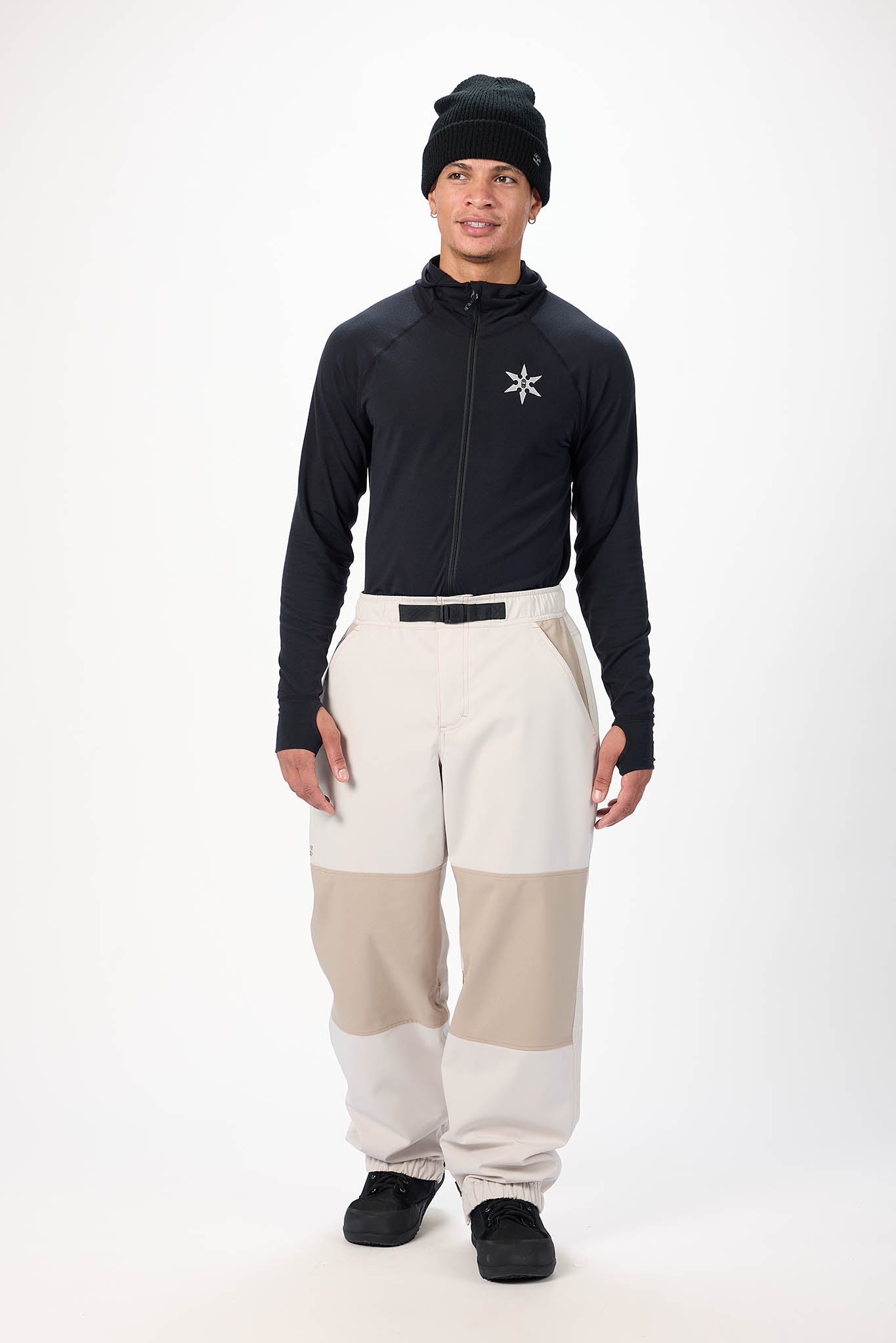 Person wearing a black jacket with a star logo, beige pants, and black beanie on a white background