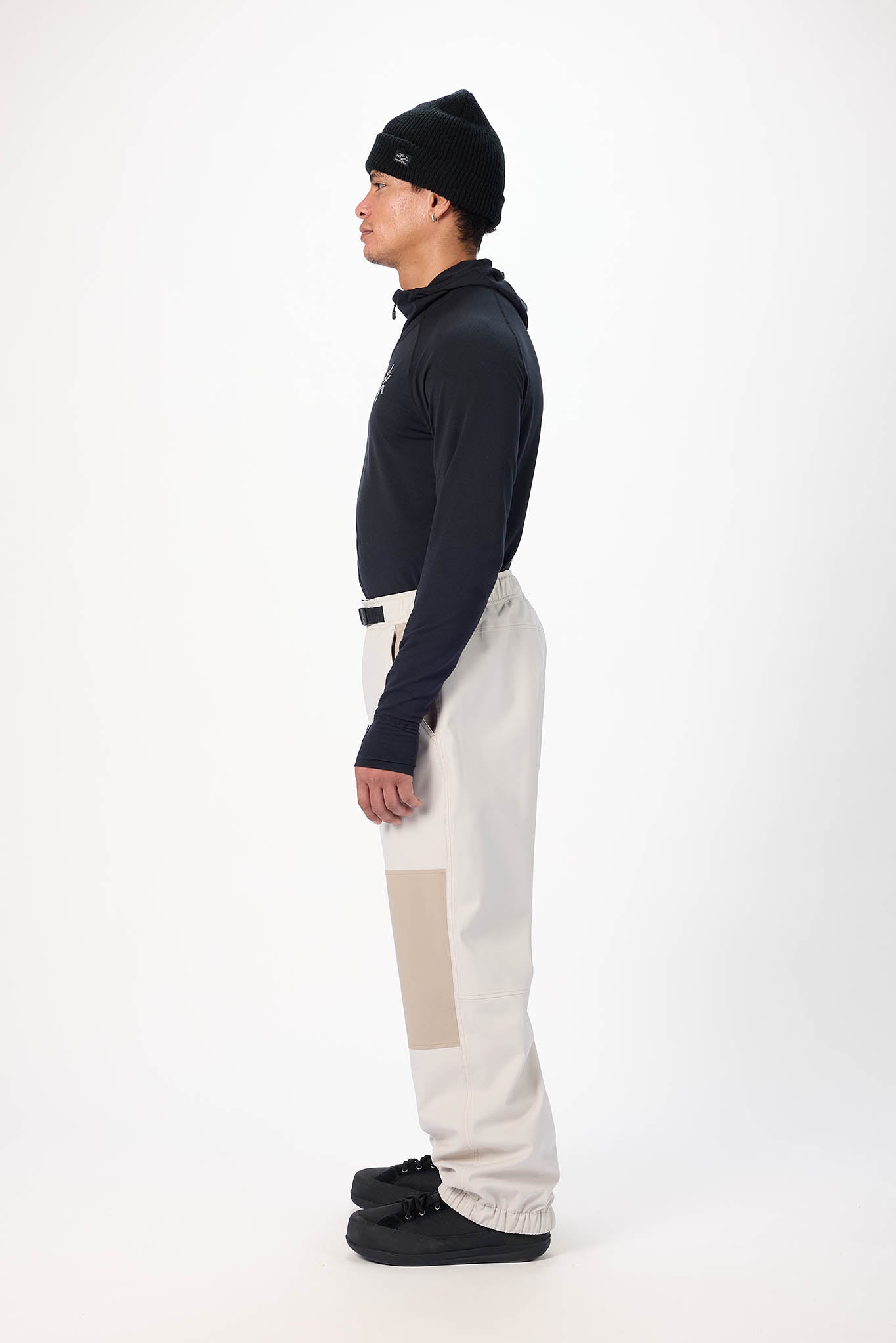 Person wearing a dark jacket, black beanie, and light-colored pants on a white background