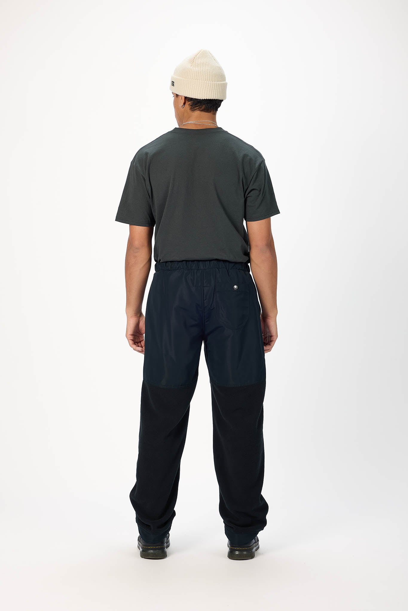 A man viewed from behind wearing a dark charcoal gray t-shirt, dark navy colorblock work pants, and a cream knit beanie against a plain white studio background.