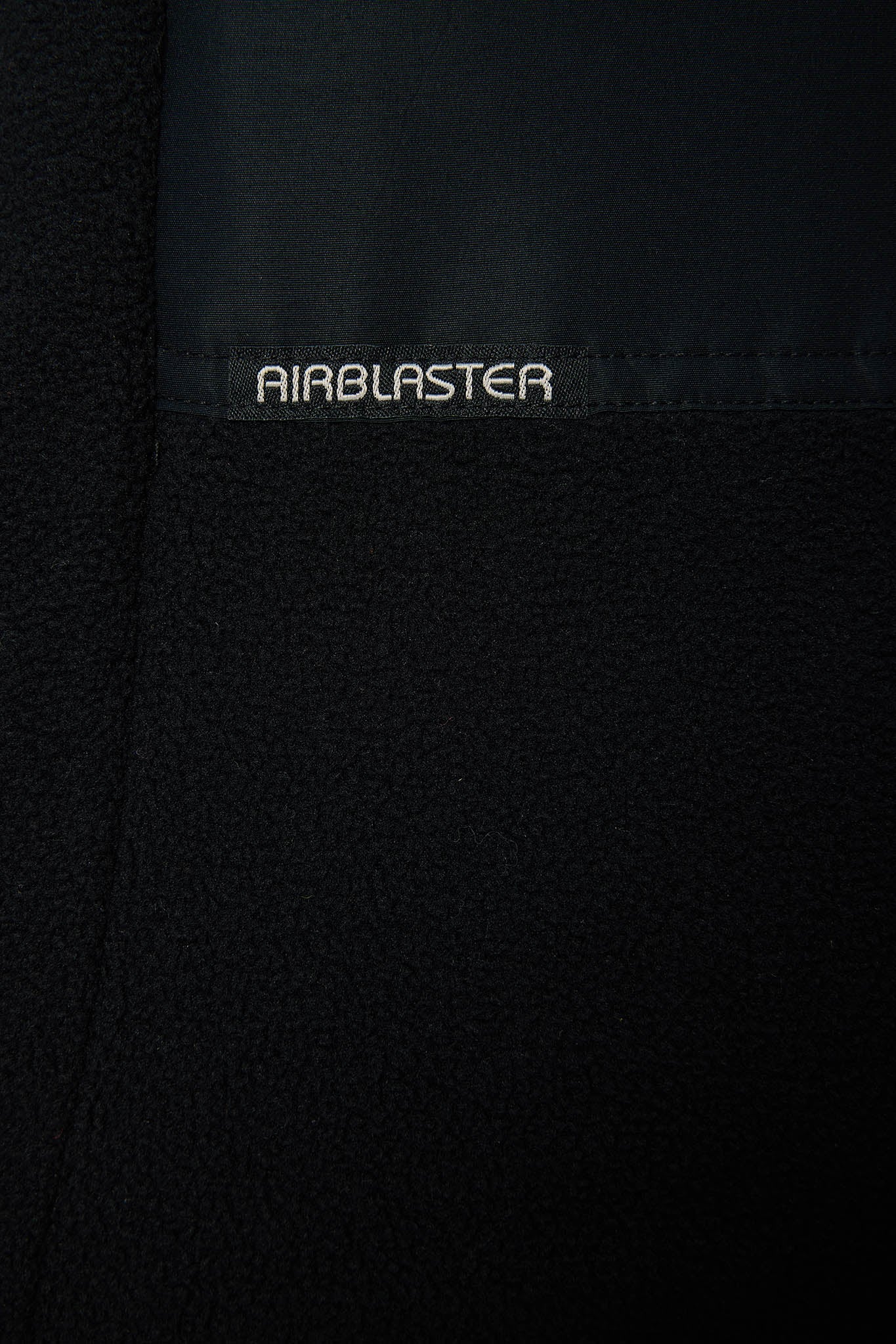 Close-up of a black textured fabric garment with a smooth black panel at the top and the word AIRBLASTER embroidered in white on the fabric.