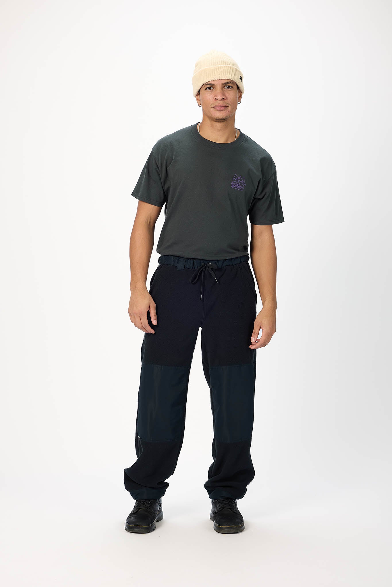 A man modeling a dark gray t-shirt with a small purple logo on the chest, paired with dark navy blue pants featuring contrast panels on the thighs, black shoes, and a cream-colored beanie, shot against a plain white studio background.