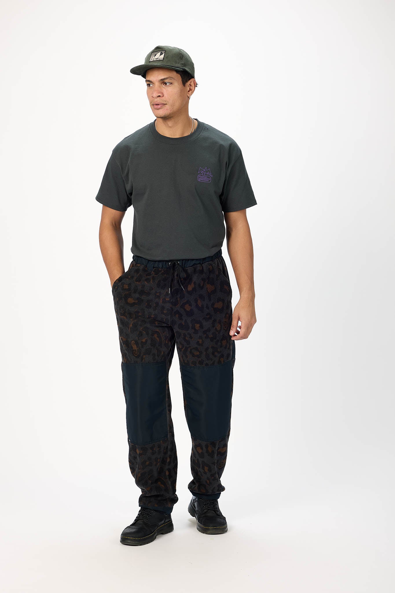 A man modeling a dark charcoal gray t-shirt with a small purple chest logo, dark patterned sweatpants with leopard-print leg patches, and a dark olive green baseball cap, standing against a plain white studio background.