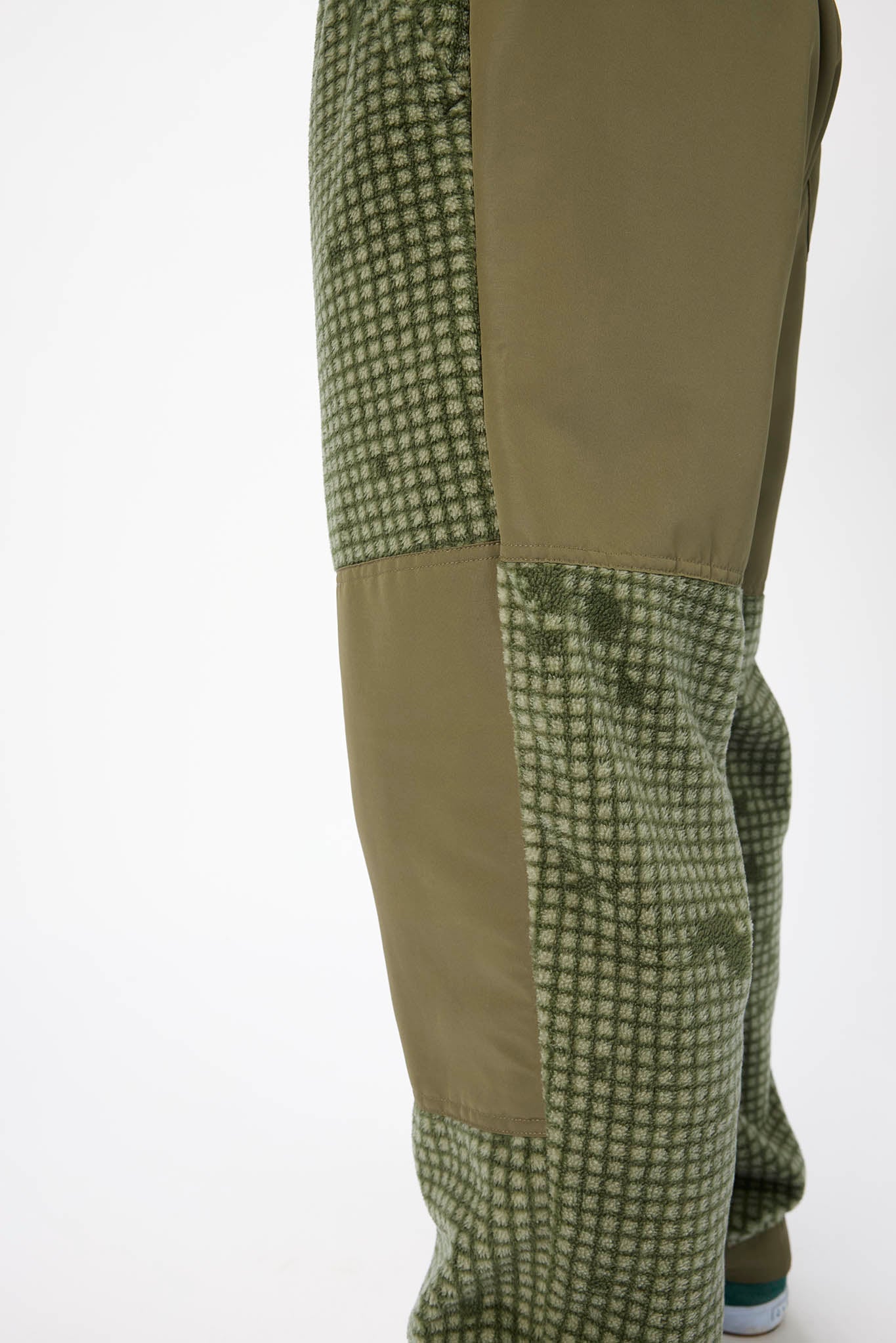 Close-up of olive green pants with a small checkered pattern on the lower legs and solid olive panels on the upper legs, shown against a plain white background.