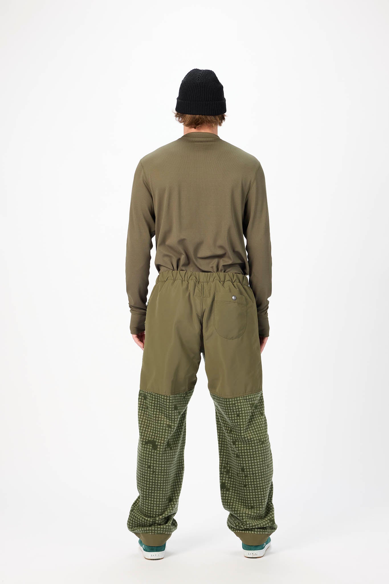 A person shown from the back wearing an olive green long-sleeve top and matching olive green pants with a darker green and black checkered pattern on the lower legs, paired with a black beanie and sneakers, against a plain white studio background.