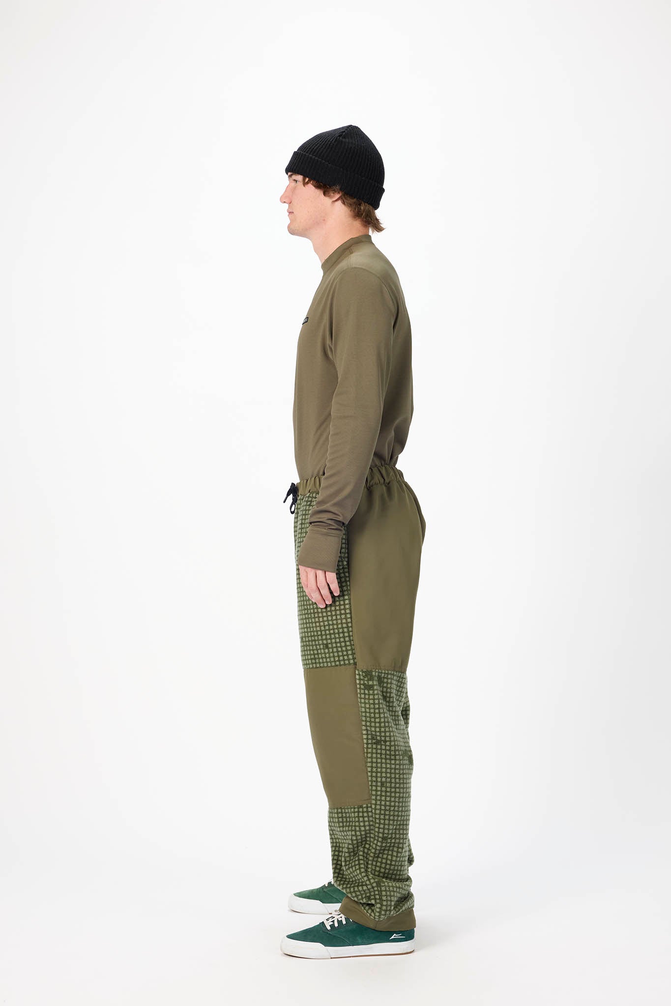 A side-profile studio shot of a model wearing an olive green long-sleeve top and matching green patchwork pants with a black beanie and green sneakers, against a plain white background.