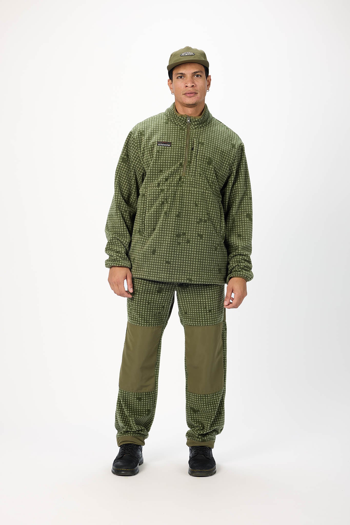 A man modeling an olive green and black checkered fleece jacket, matching pants, and a green cap against a plain white studio background.
