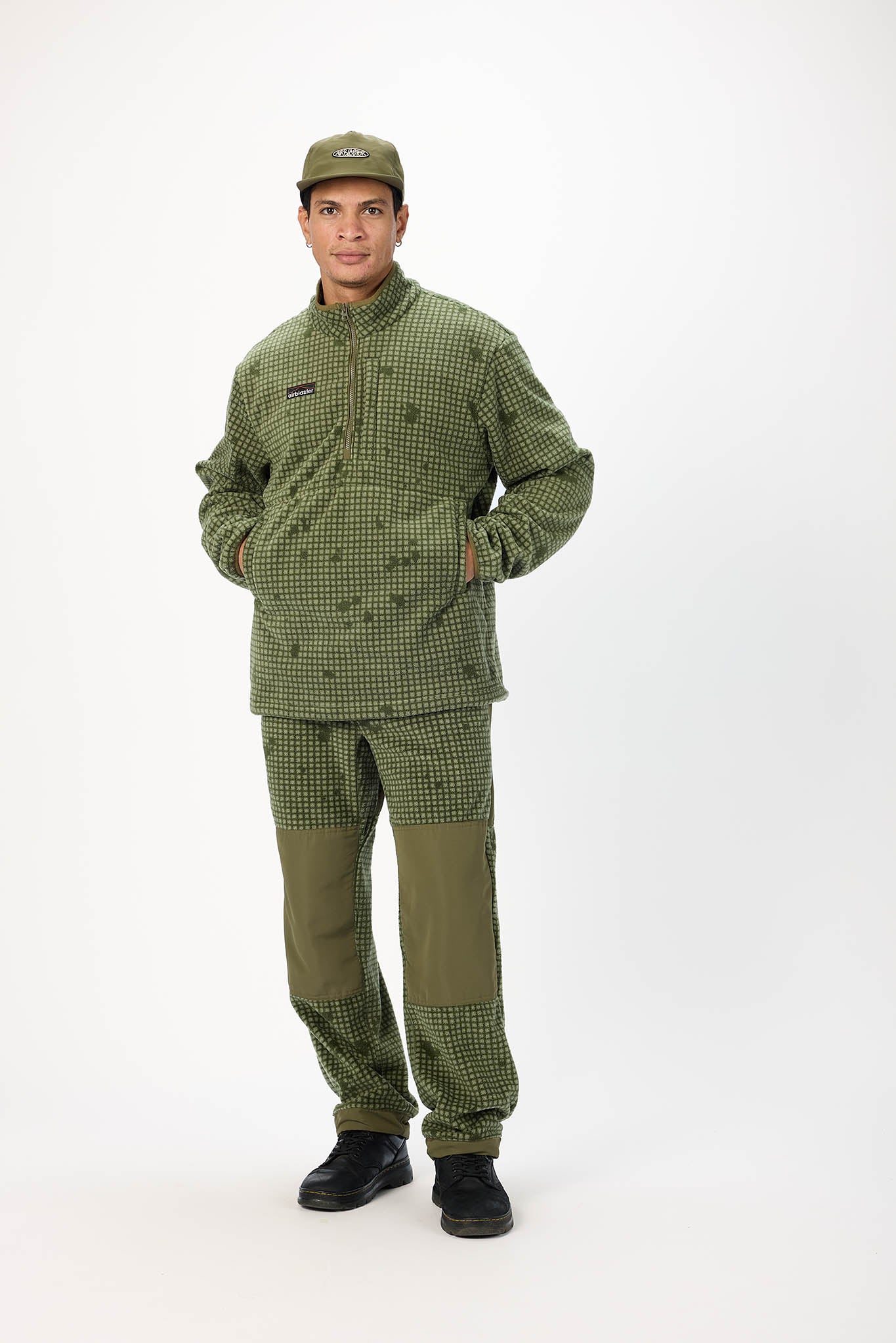 A man modeling an olive green work uniform set consisting of a long-sleeve button-up jacket, matching pants with solid olive green knee patches, and an olive green baseball cap, shot against a plain white studio background.