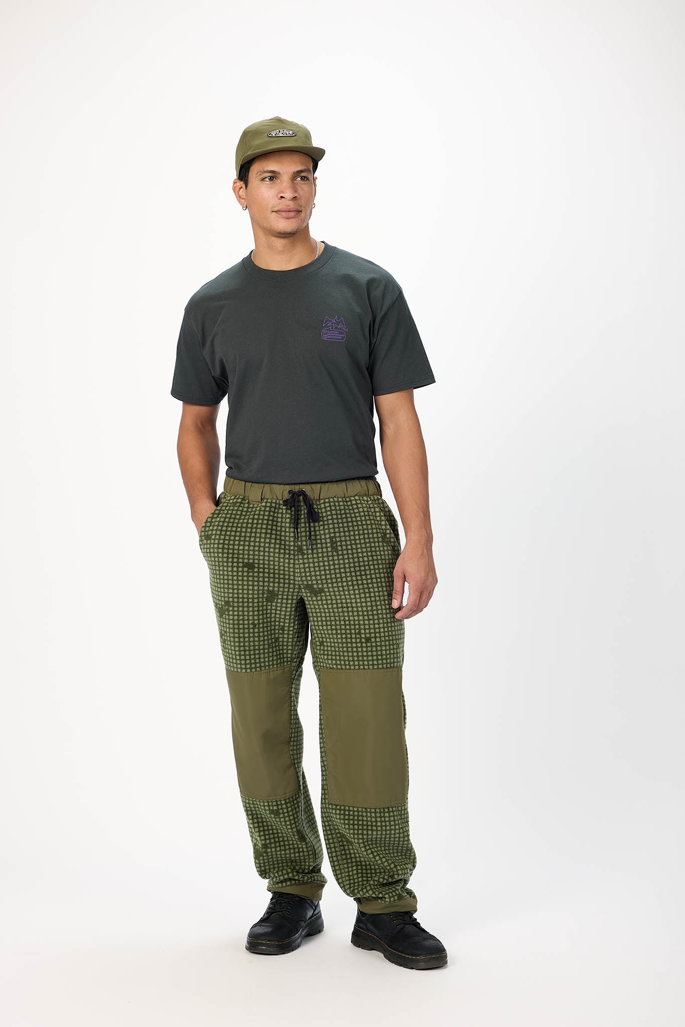 A man modeling a dark charcoal gray t-shirt, olive green and black checkered cargo pants with solid olive green knee patches, and an olive green baseball cap, standing against a plain white studio background.