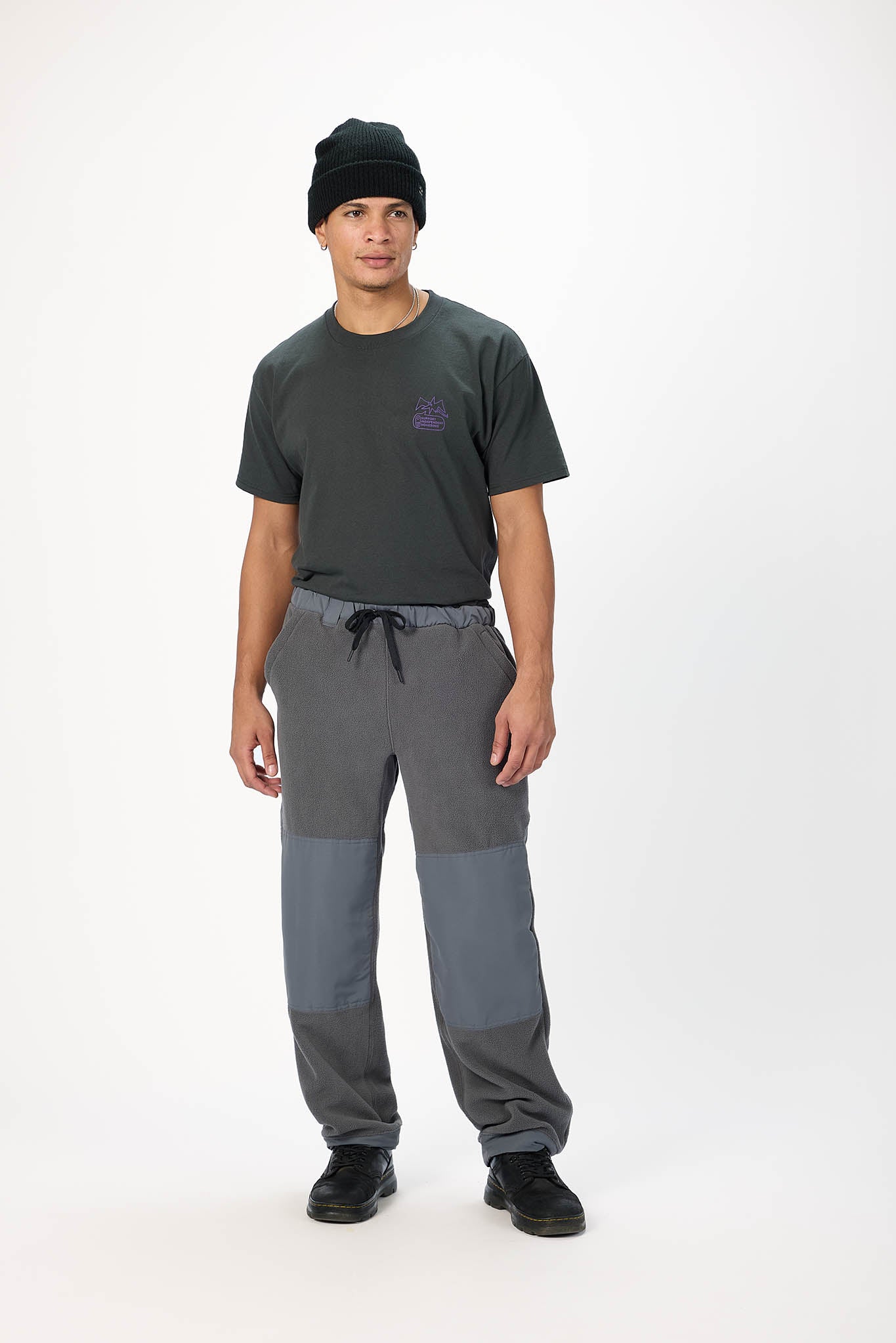 A man modeling a dark gray t-shirt, gray and blue color-blocked pants, and a black beanie against a plain white studio background.