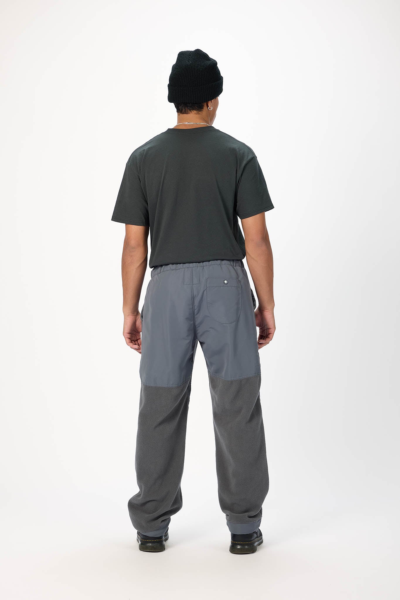 A man viewed from behind wearing a dark charcoal t-shirt, gray and slate blue colorblock cargo pants, and a black beanie against a plain white studio background.