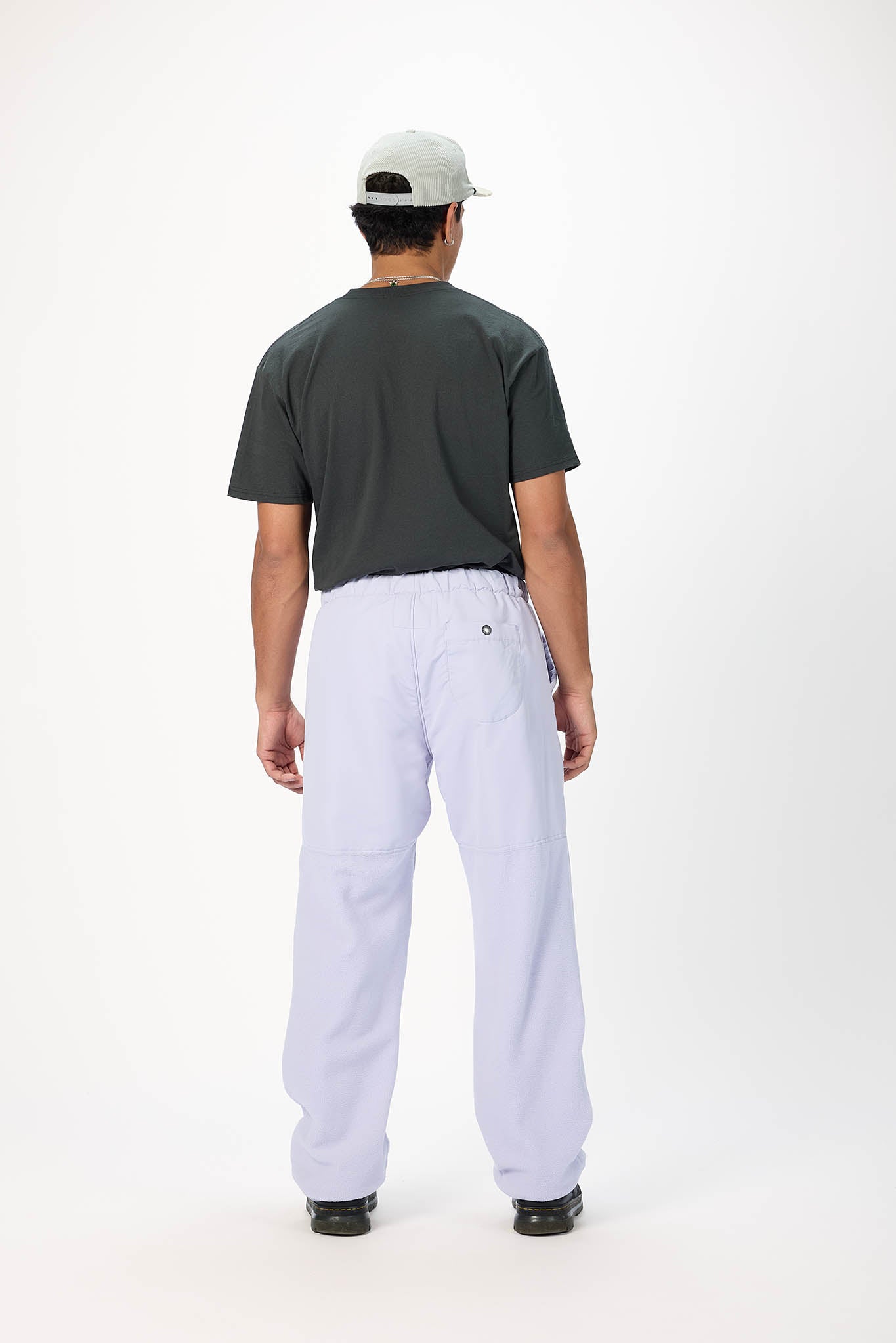 A man viewed from behind wearing a dark charcoal t-shirt, light lavender pants, and a light gray baseball cap against a plain white studio background.