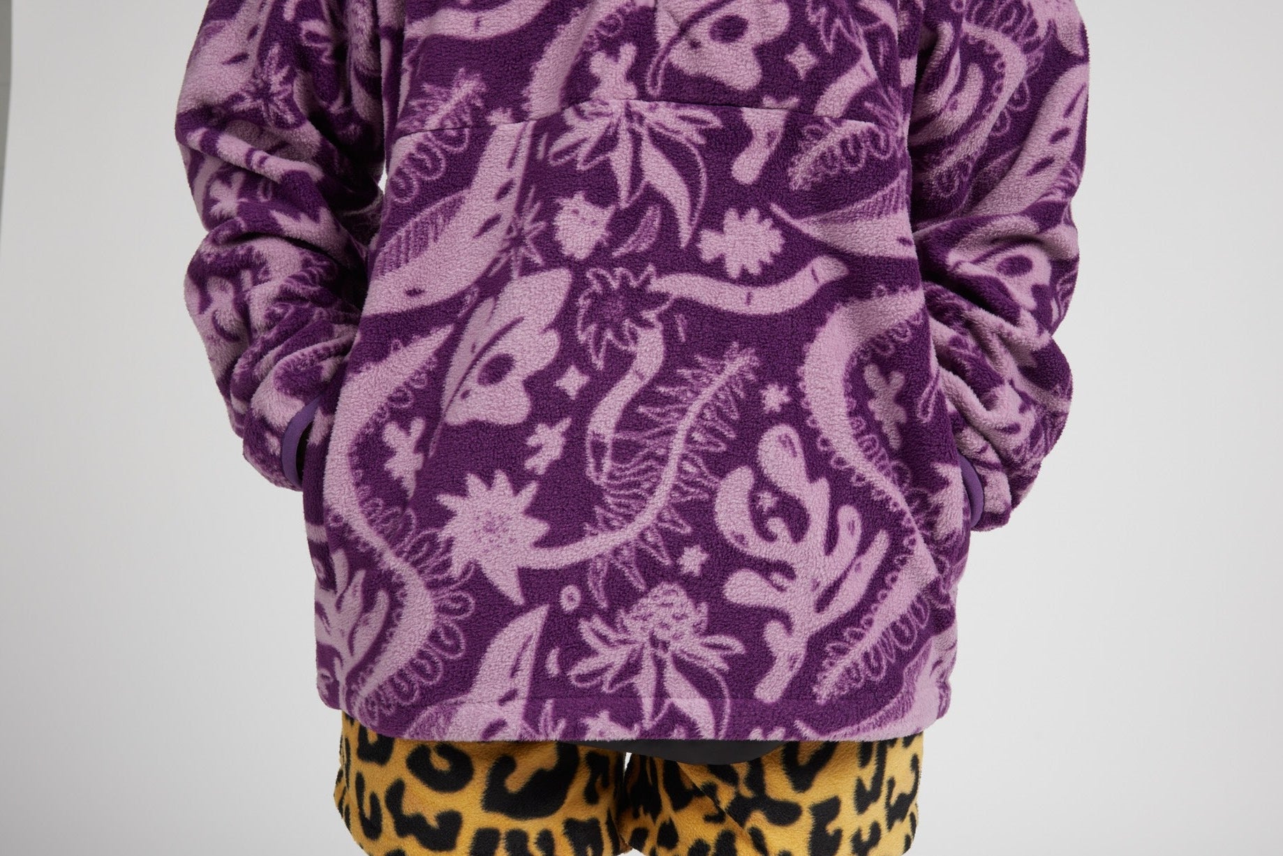 A person wearing a purple fleece pullover with a light purple floral and abstract pattern, paired with yellow leopard-print shorts, photographed against a light gray studio background.