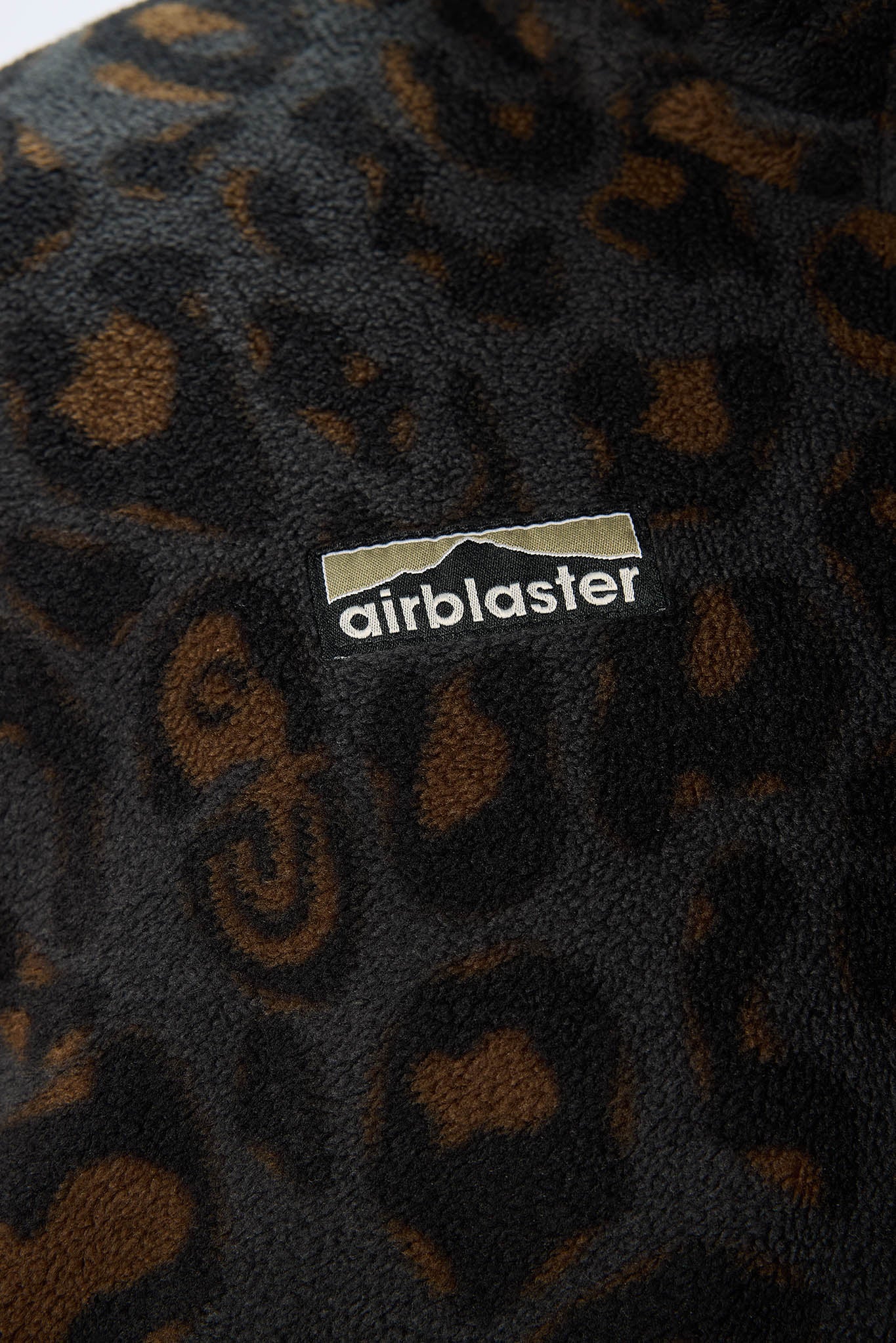 Close-up of a plush fleece fabric with a dark leopard print pattern in black and brown, featuring a white and gray Airblaster logo patch.