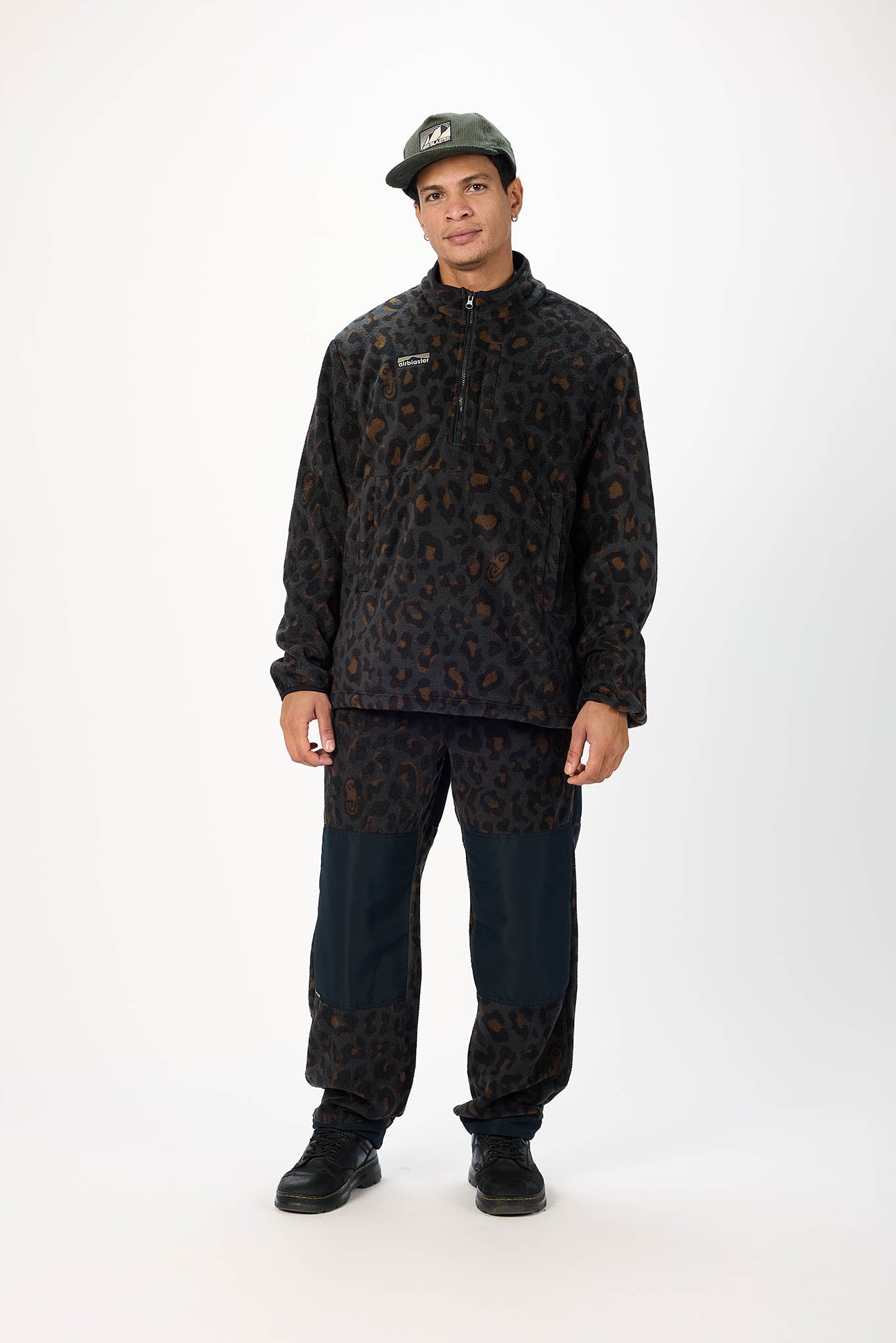 A man modeling a dark patterned quarter-zip fleece pullover, matching pants, and a dark cap against a plain white studio background.