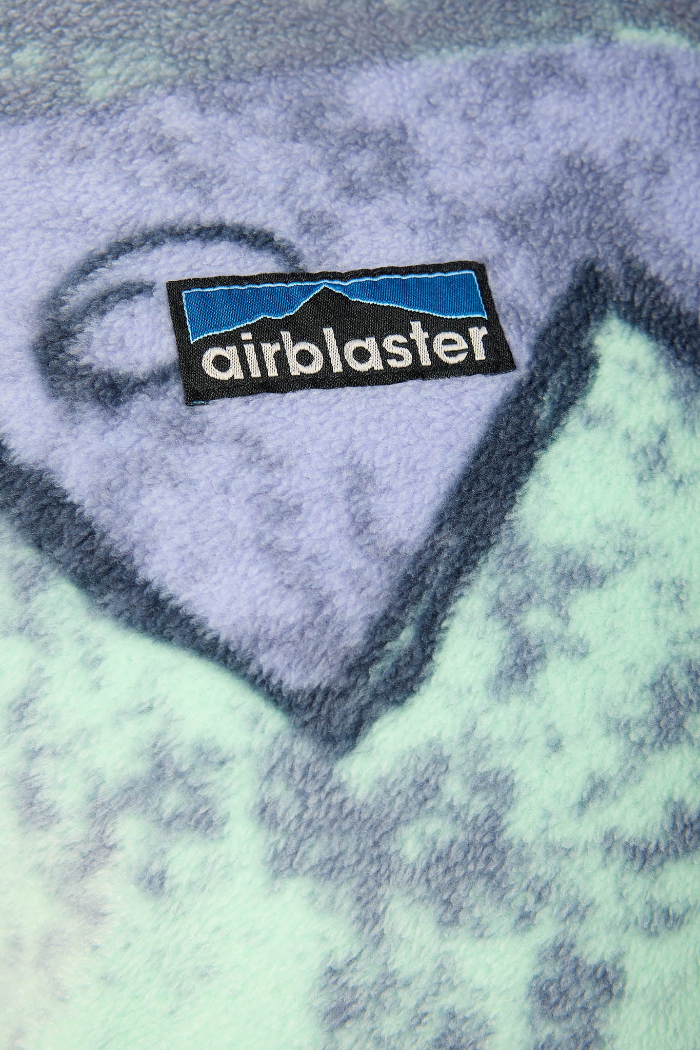 Close-up of a fuzzy fleece blanket with a heart-shaped pattern in lavender and mint green, featuring a black embroidered patch with the Airblaster logo.