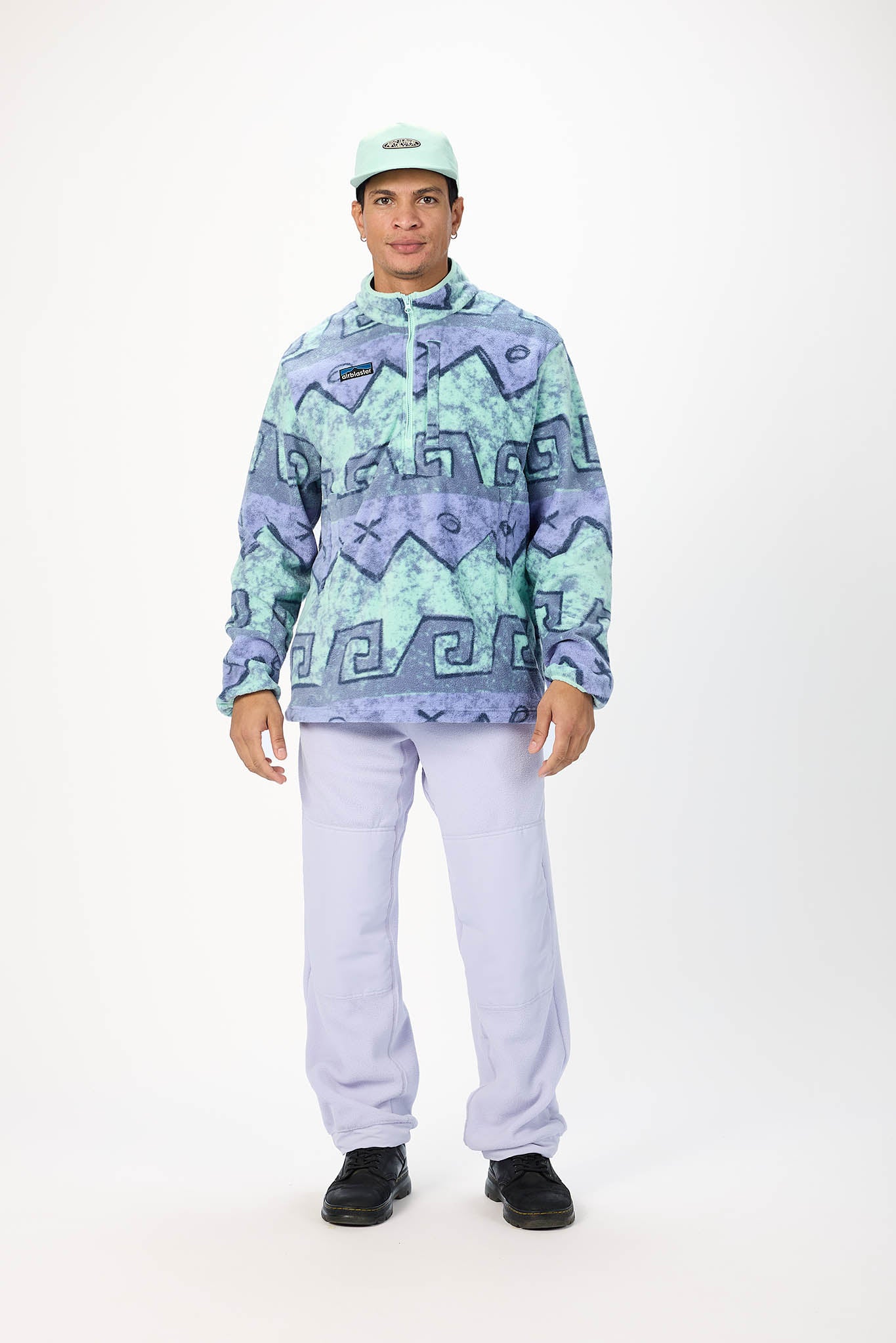 A model wearing a light green half-zip pullover fleece with a blue and green geometric pattern, paired with light purple sweatpants, black sneakers, and a light green cap, standing against a white studio background.