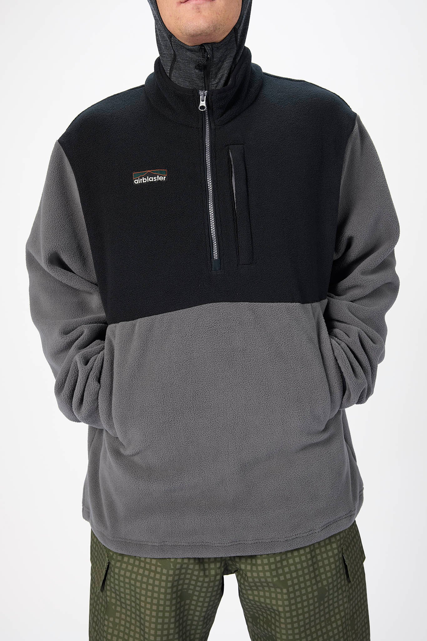 A man wearing a quarter-zip fleece pullover with a black upper chest and shoulders and a gray lower body and sleeves, paired with olive green patterned pants, against a plain white background.