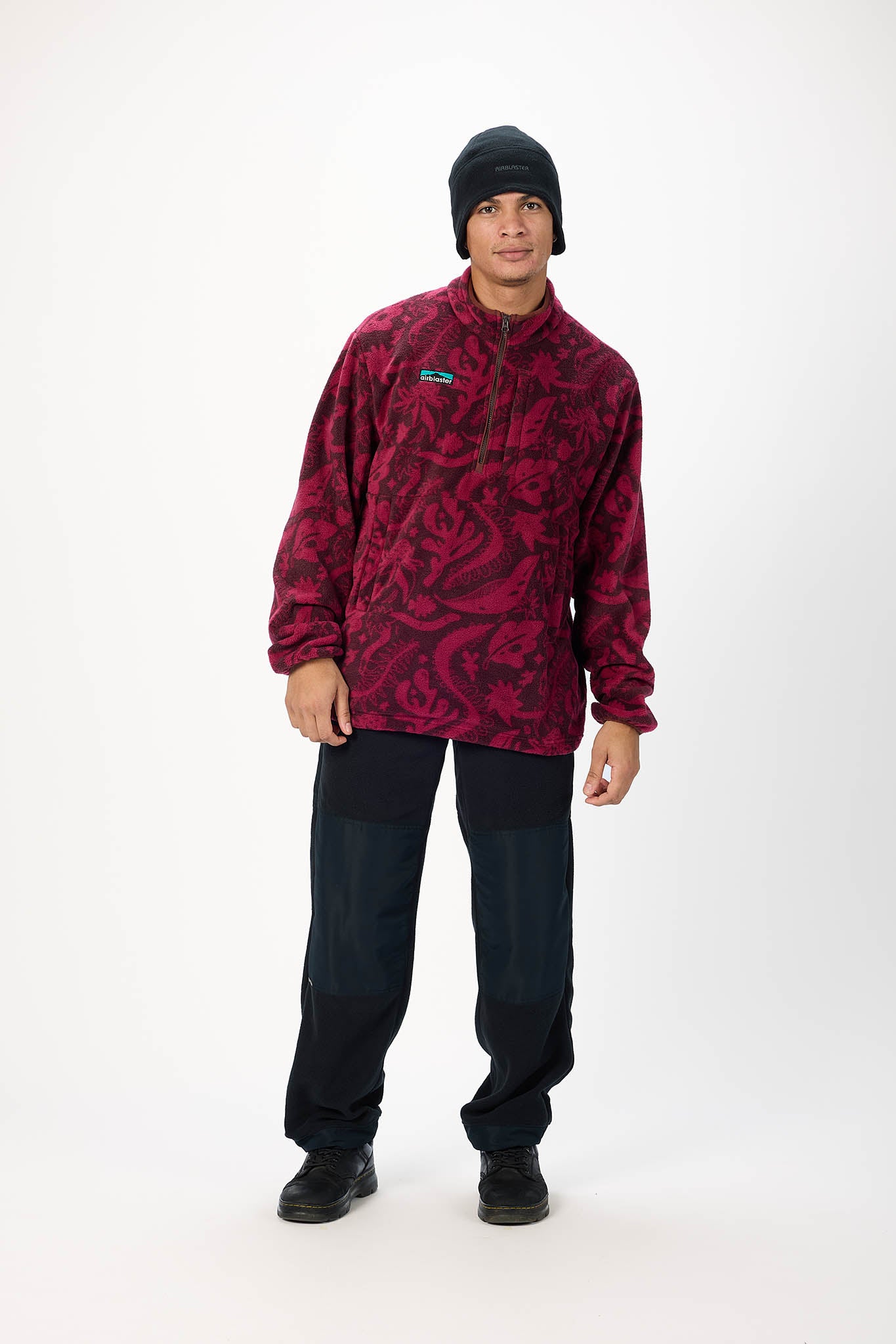 A man modeling a quarter-zip fleece pullover in a dark red and black paisley pattern, paired with dark pants and a black beanie, against a plain white studio background.
