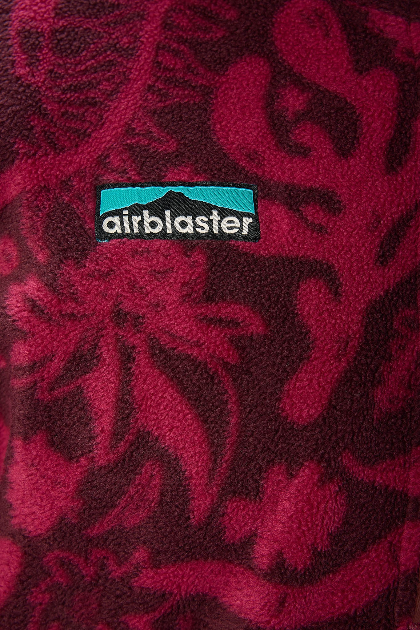 A close-up of a plush fleece fabric with a dark red and magenta floral pattern, featuring a central black Airblaster logo patch with a teal background.