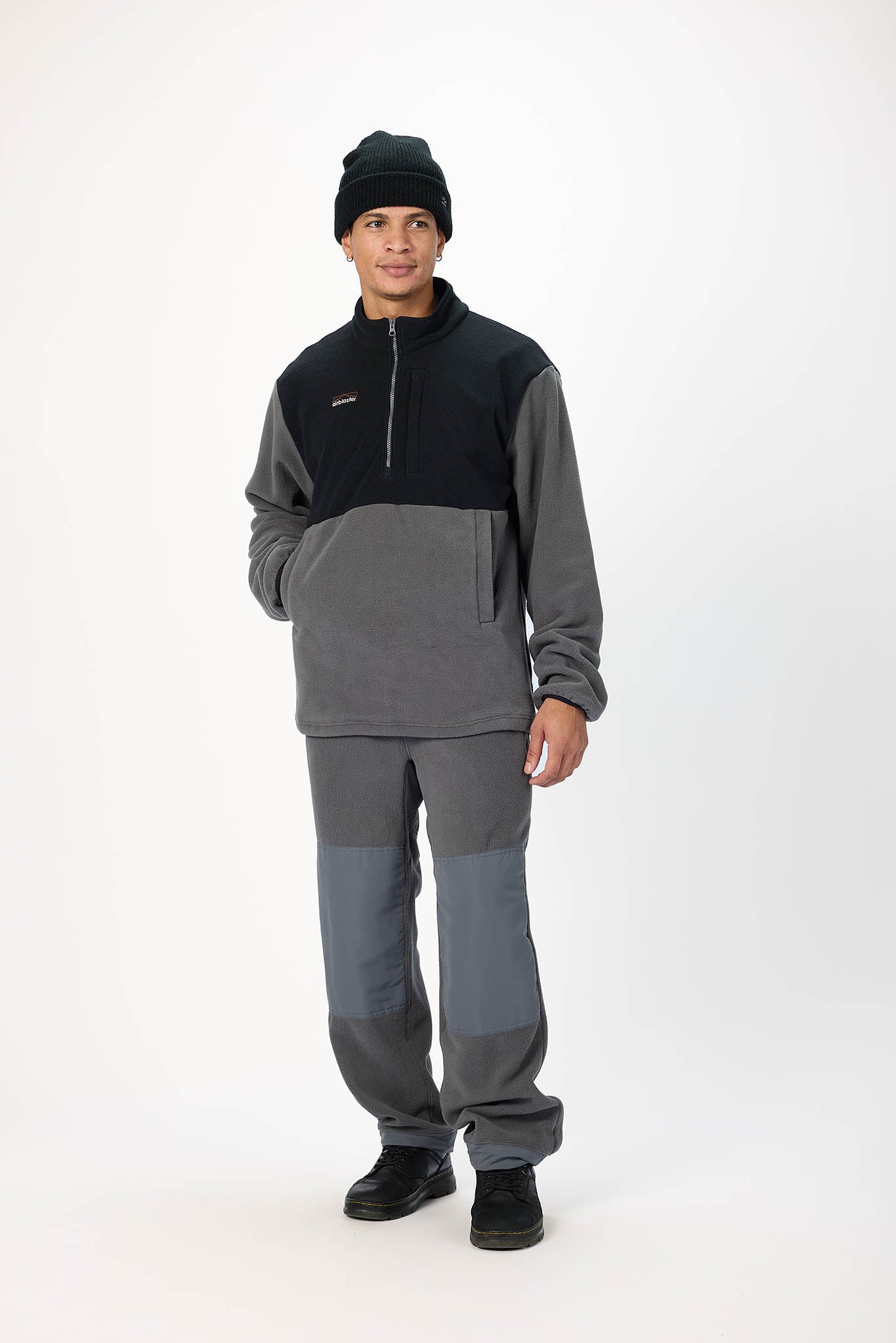 A man modeling a two-tone gray and black fleece quarter-zip pullover and matching gray pants with black shoes and a black beanie, photographed against a plain white studio background.