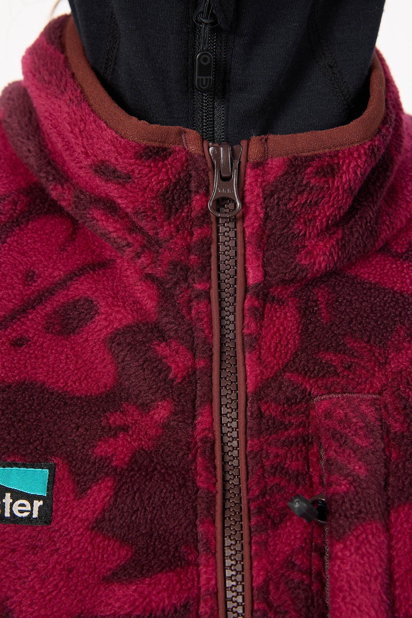Close-up of a fleece jacket with a dark red and black camouflage pattern, a brown collar, and a black zipper. A small black and teal Patagonia logo patch is visible on the lower left.