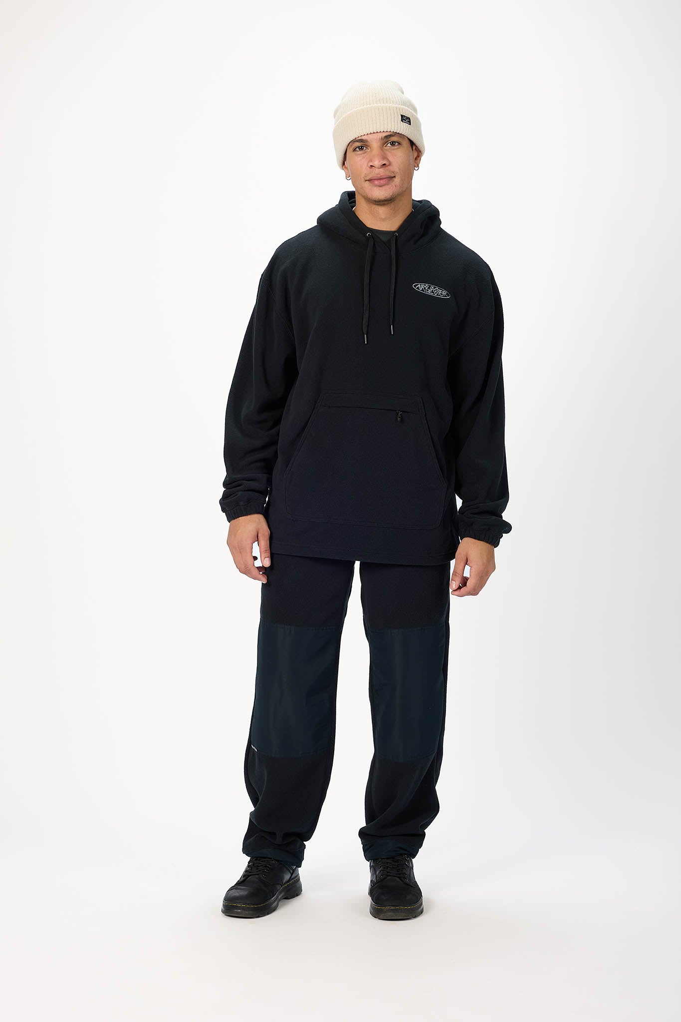 A man modeling a black pullover hoodie with a small white logo on the left chest, paired with dark pants, black shoes, and a cream beanie, standing against a plain white studio background.
